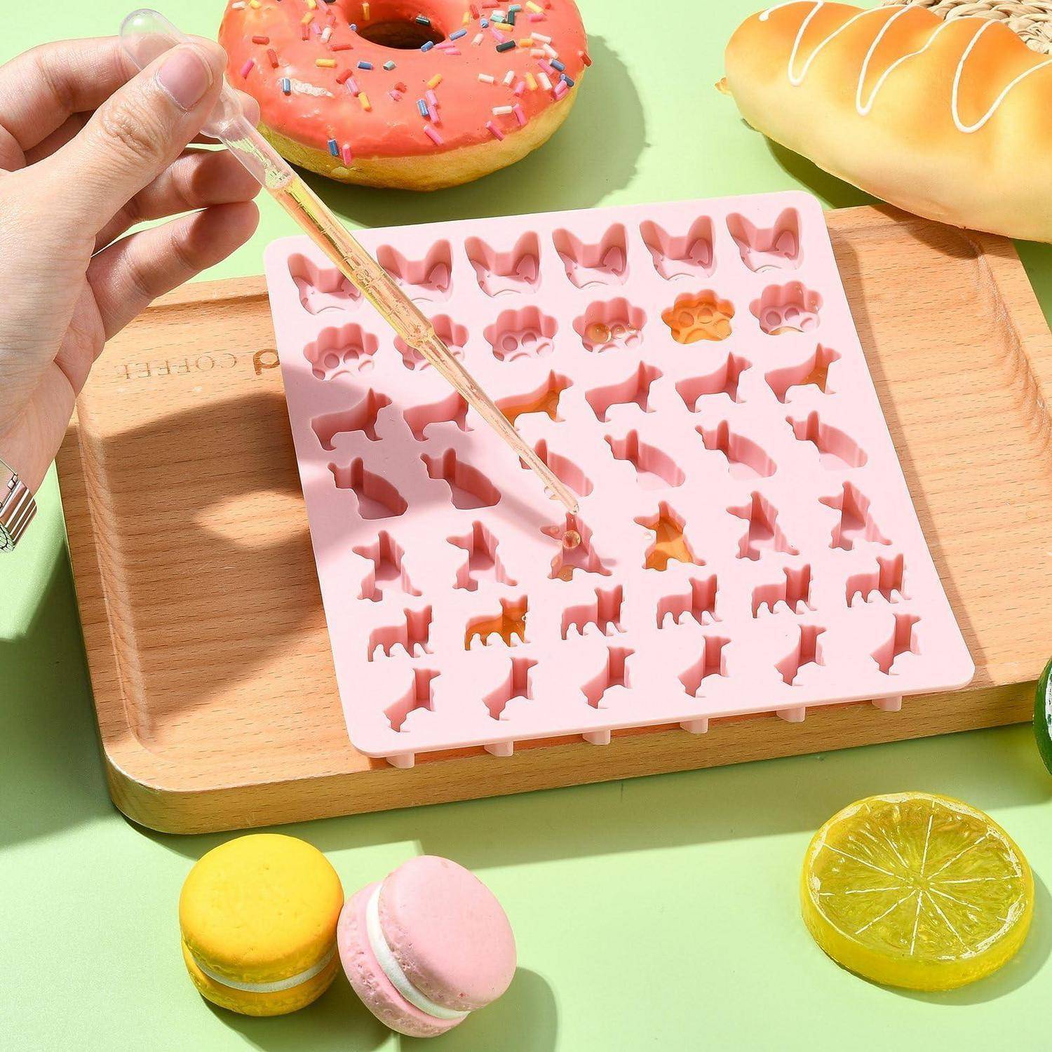Eine Hand hält ein dünnes Werkzeug über eine rosa Silikonform, die mit Tier- und Formhohlräumen gefüllt ist, auf einem Holztablett, mit bunten Gebäcken darum herum.