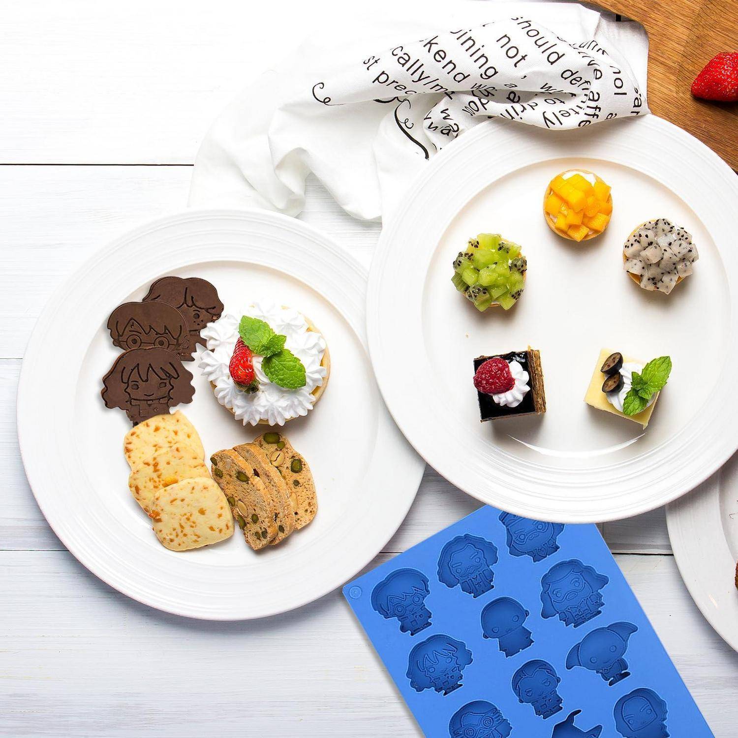 Teller mit sortierten dekorierten Keksen und Petit Fours, mit einem blauen Silikonform-Tablett und einem weißen Tuch im Hintergrund.