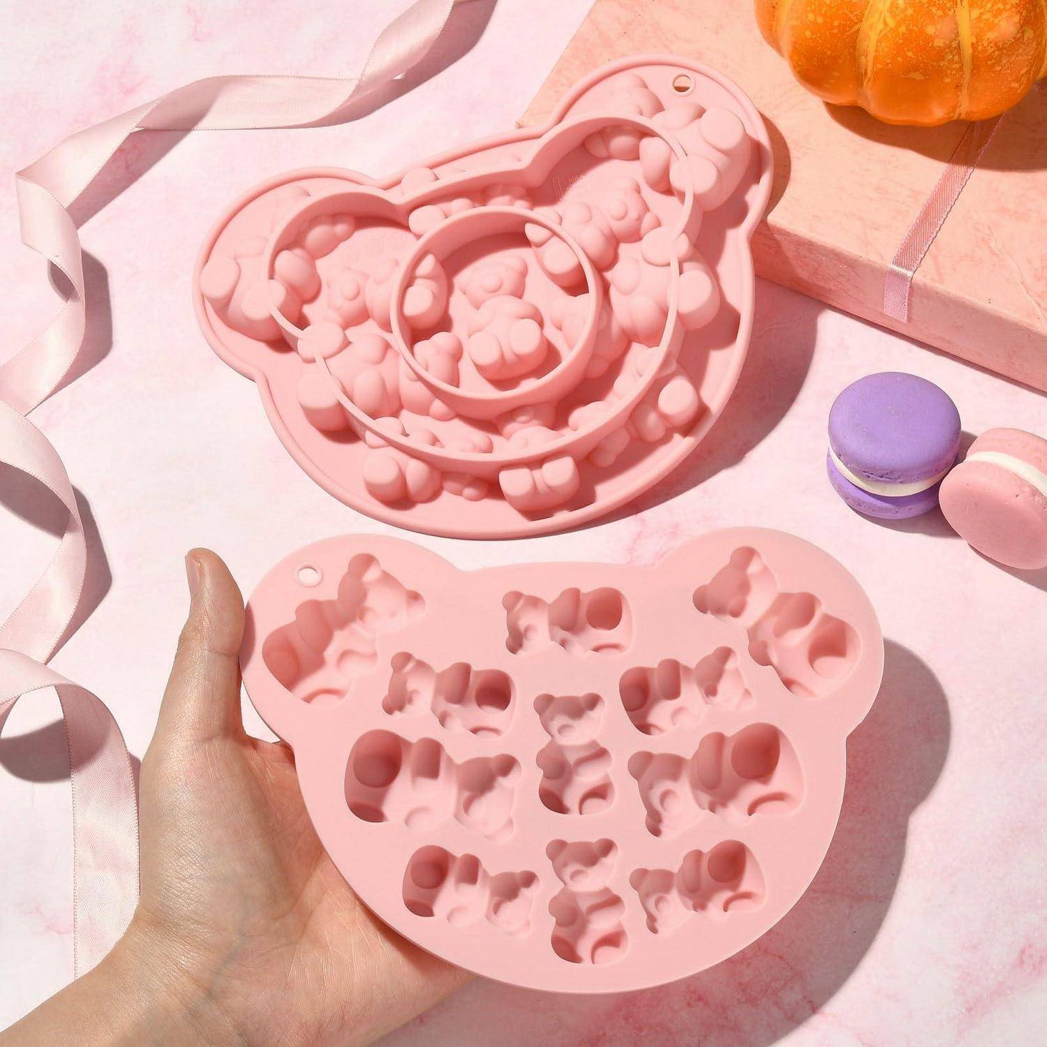 Hand hält eine rosa teddybärförmige Silikonform neben einem passenden Tablett mit kleinen Tier- und Herz-Vertiefungen, mit pastellfarbenen Makronen und einem Kürbis im Hintergrund.