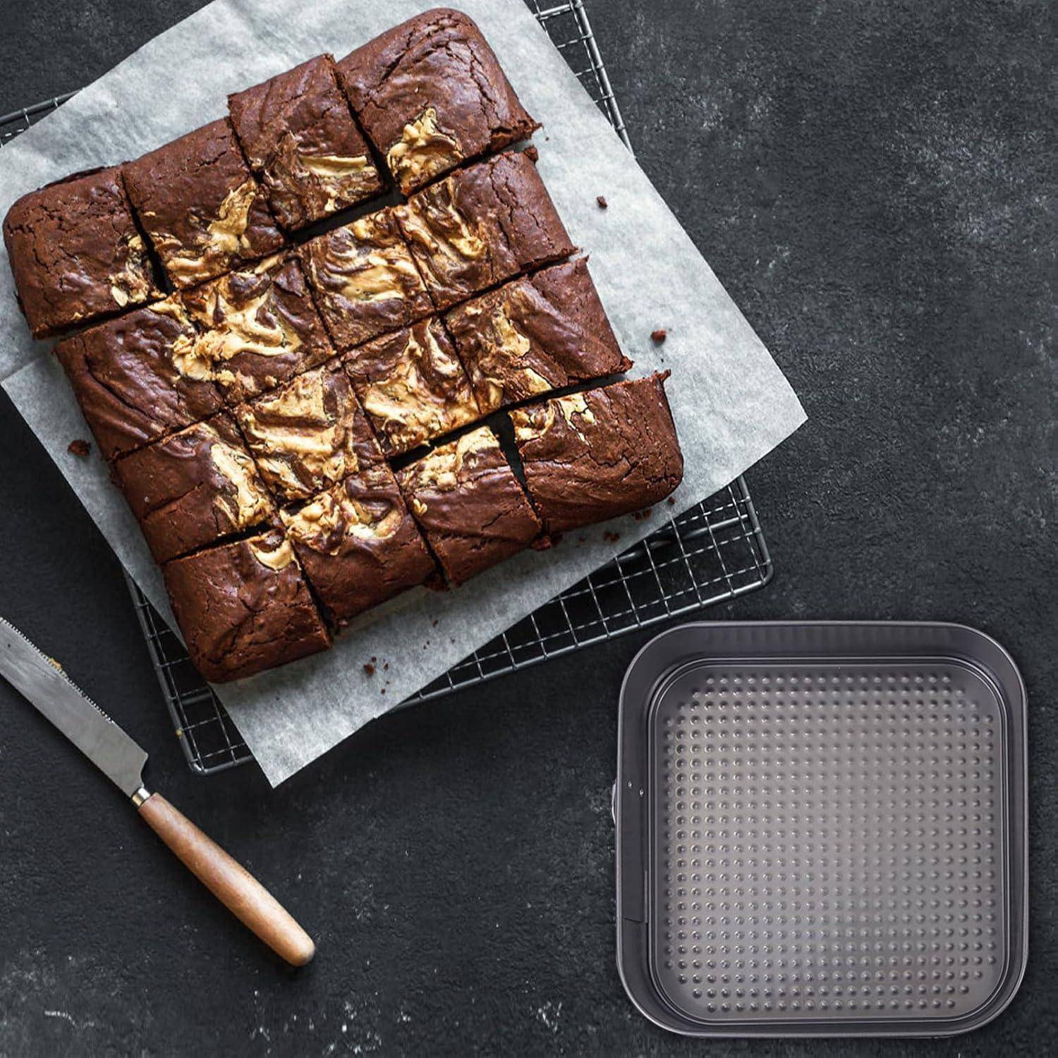 Quadratische Schokoladenbrownie, in 16 Stücke geschnitten, auf Backpapier über einem Kühlgitter, mit einem Messer und einer leeren Backform in der Nähe.