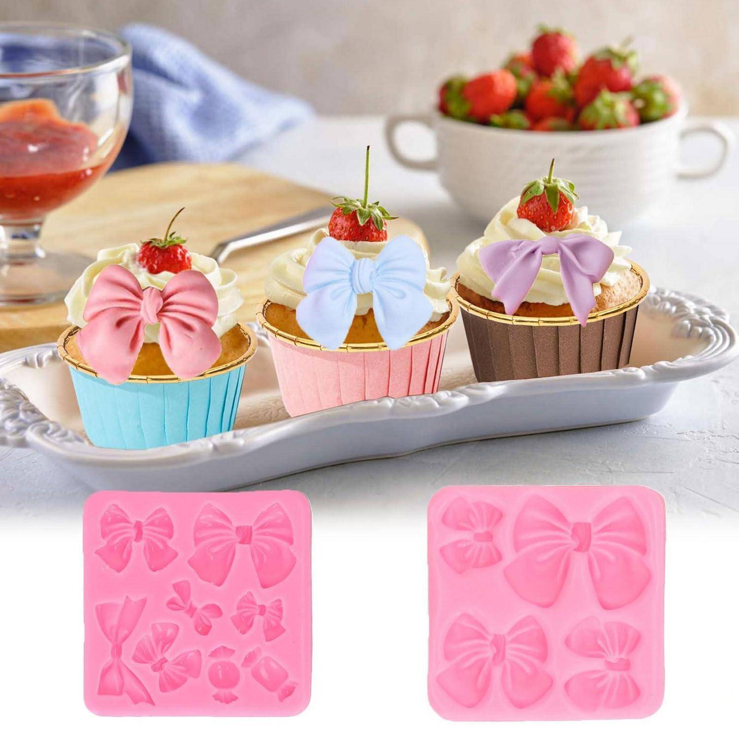 Dekorierte Erdbeercupcakes auf einem weißen Tablett, dazu zwei rosa Silikonform-Tabletts mit Schleifen-Formen zum Herstellen von essbaren Dekorationen.