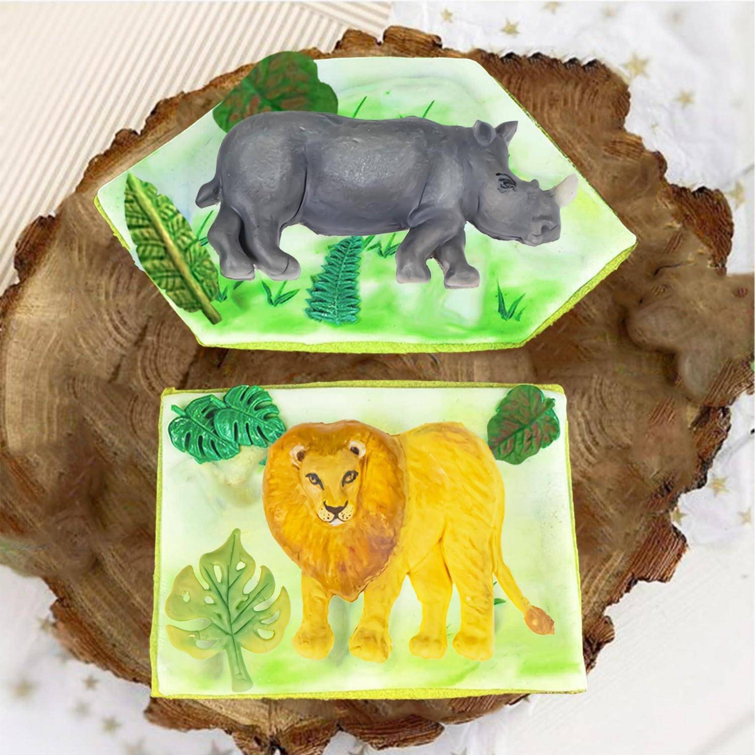 Zwei Fondant-Tierszenen auf einer Holzscheibe: ein grauer Nashorn oben und ein gelber Löwe unten, beide auf grünen blättrigen Sockeln.
