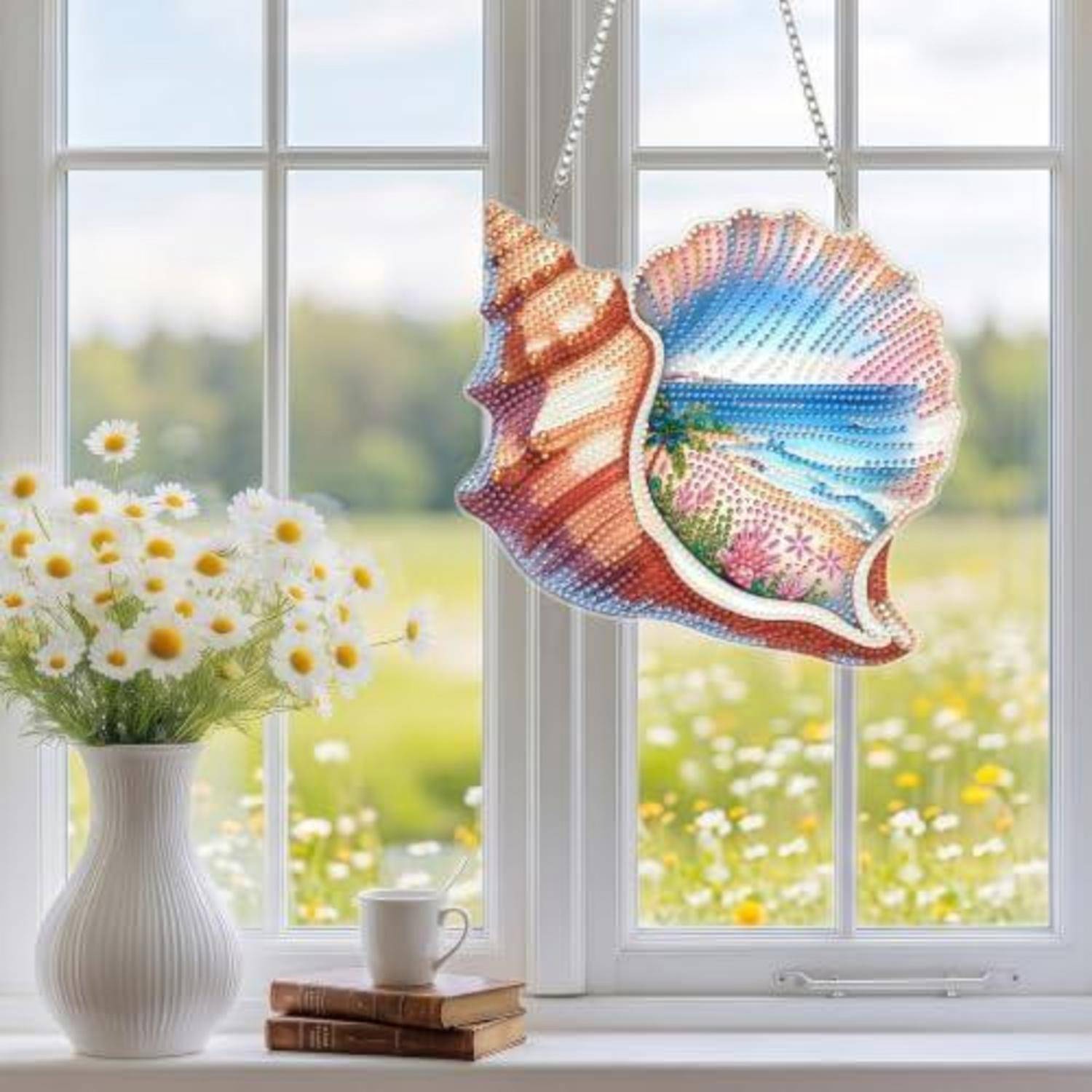 Ein bunter Muschel-Sonnenfänger hängt in einem hellen Fenster neben einer Vase mit Gänseblümchen, einer Tasse und Büchern auf der Fensterbank.