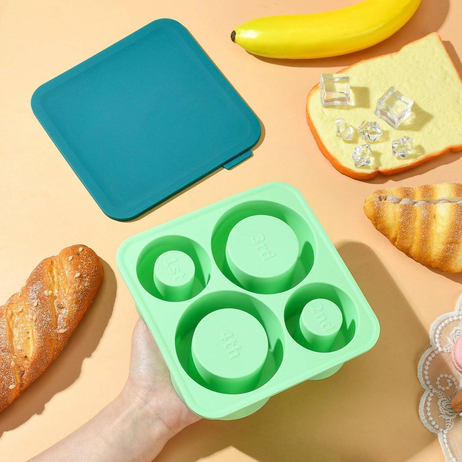Hand, die einen minzgrünen vierteiligen Behälter mit runden Einsätzen hält, umgeben von Brot, Banane und juwelenähnlichen Stücken auf pastellfarbenem Hintergrund.