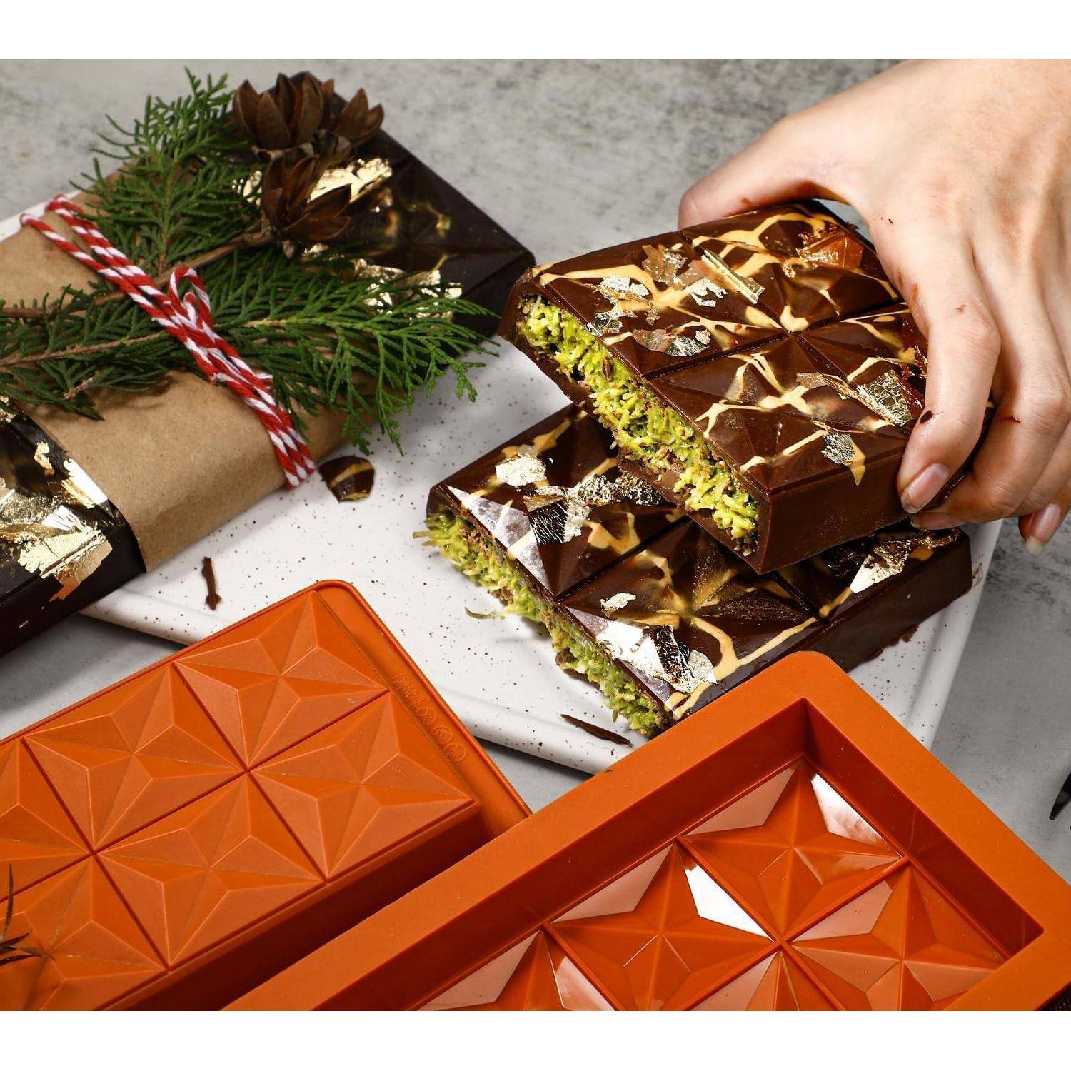 Hand öffnet eine geschichtete Schokolade mit Pistazienfüllung auf einem Teller, daneben eingewickelte Weihnachtssüßigkeiten und orange Geschenkboxen.