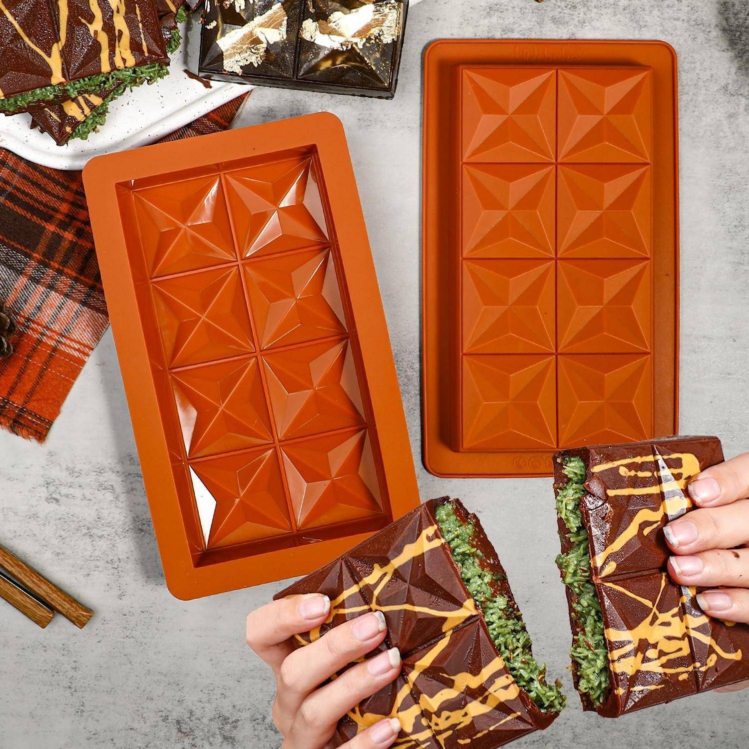 Hände halten zwei Schokoladentafeln mit grüner Füllung neben orangefarbenen geometrischen Schokoladenformen und verschiedenen Schokoladenstücken auf einer Tischplatte.