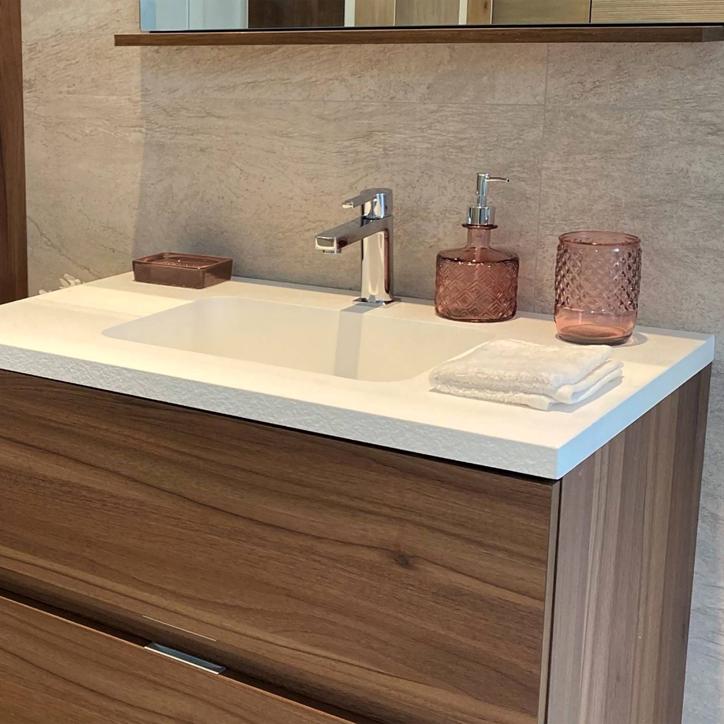 Modernes Badezimmerwaschtisch mit weißem Waschbecken, Chromarmatur, rosa Seifenspender und Zahnputzbecher, zusammengefaltete Handtücher und Unterschrank mit Holzfront.