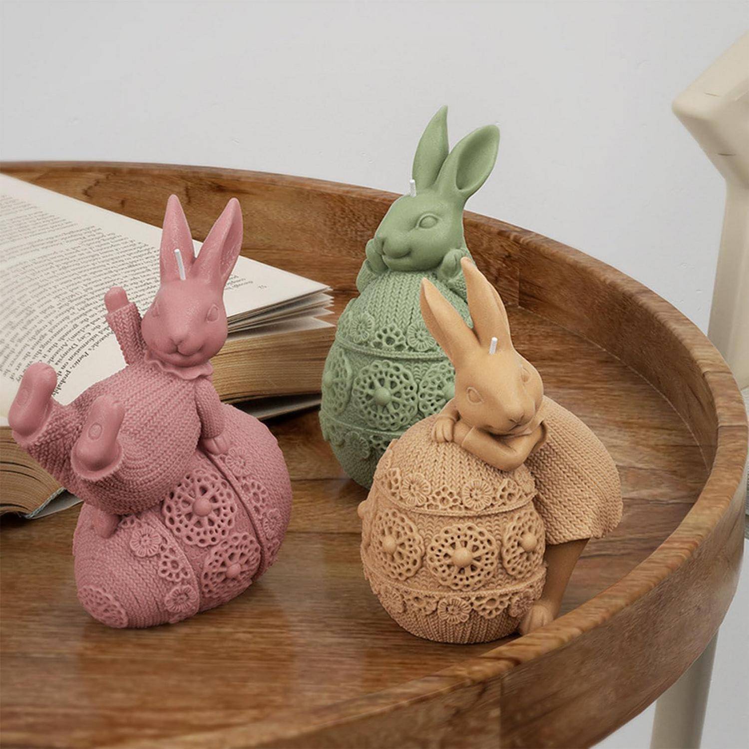Drei dekorative gestrickte Häschenfiguren in Rosa, Grün und Beige sitzen auf einem Holztablett neben einem aufgeschlagenen Buch.