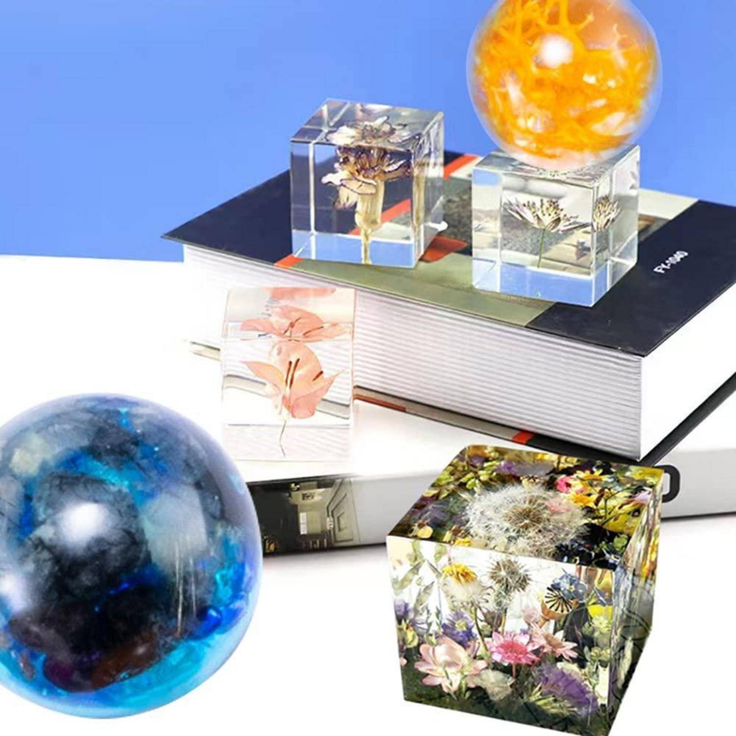 Dekorative Kunstharz-Würfel und -Kugeln mit konservierten Blumen und Insekten, angeordnet auf Büchern, mit einer großen blauen Kugel im Vordergrund.