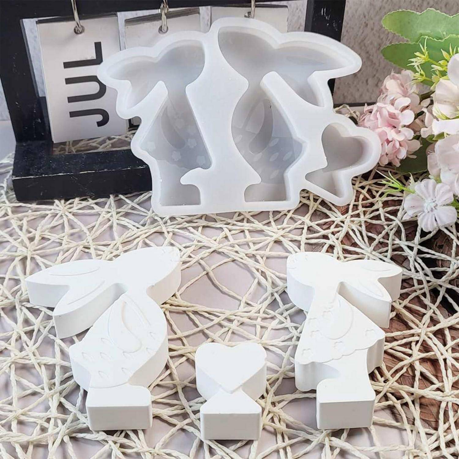 Weiße Silikonform mit Herz- und Flügelformen und vier kleine fertige weiße Dekorationsstücke, die auf einer geflochtenen Matte angeordnet sind, mit Blumen im Hintergrund.