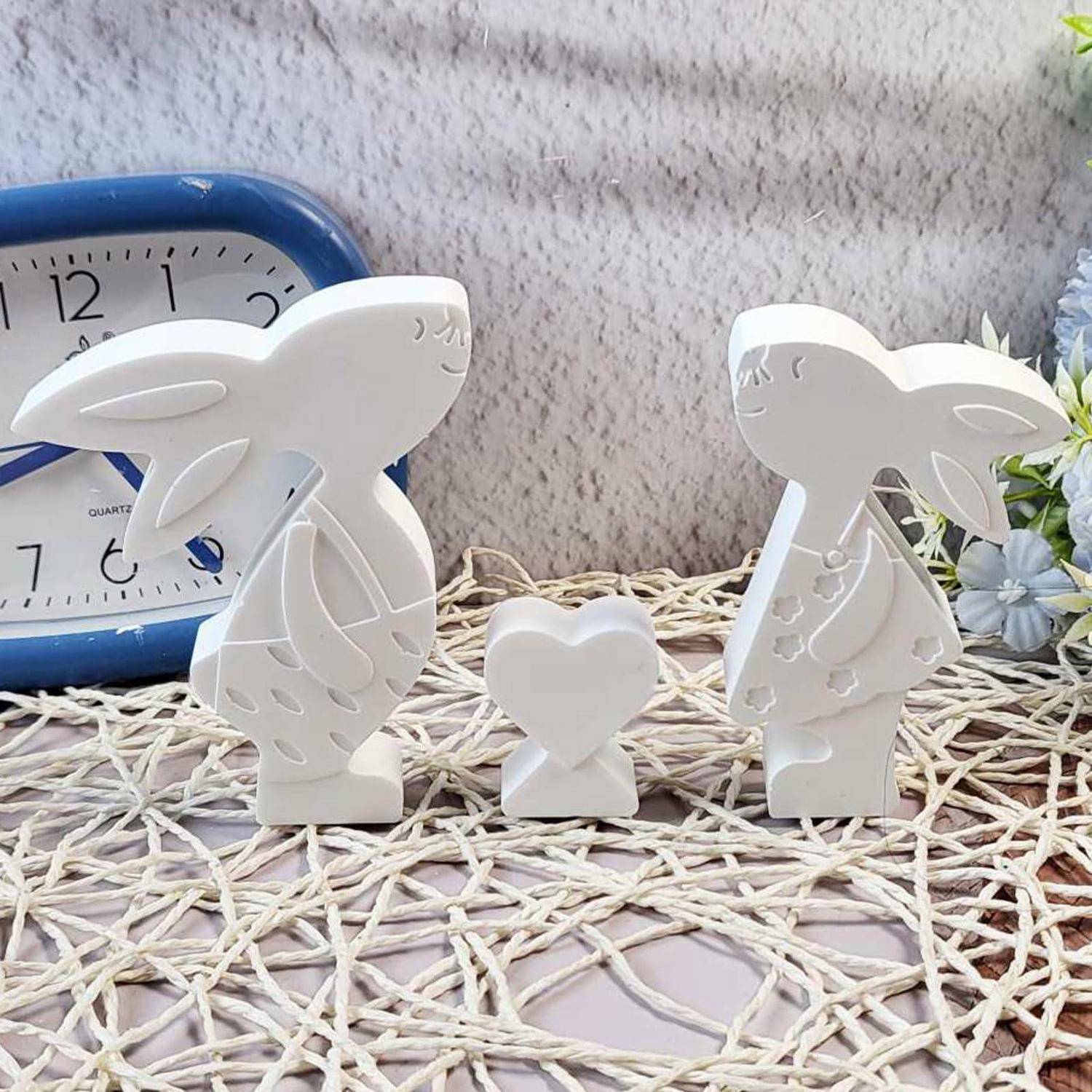 Zwei weiße Kaninchenfigurinen schauen sich gegenseitig an, mit einem kleinen Herz zwischen ihnen, ausgestellt auf einer Webmatte neben einer blauen Uhr und Blumen.
