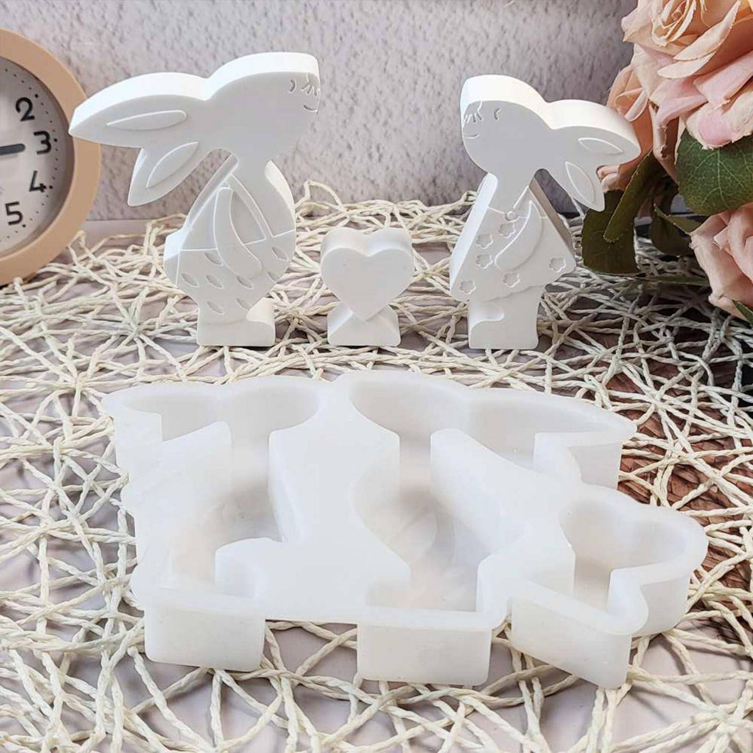 Weiße 3D-gedruckte Kaninchen- und Herzdekorationen, die über einer großen kaninchenförmigen Form oder einem Puzzleteil auf einer geflochtenen Matte angezeigt werden, mit Blumen und einer Uhr in der Nähe.