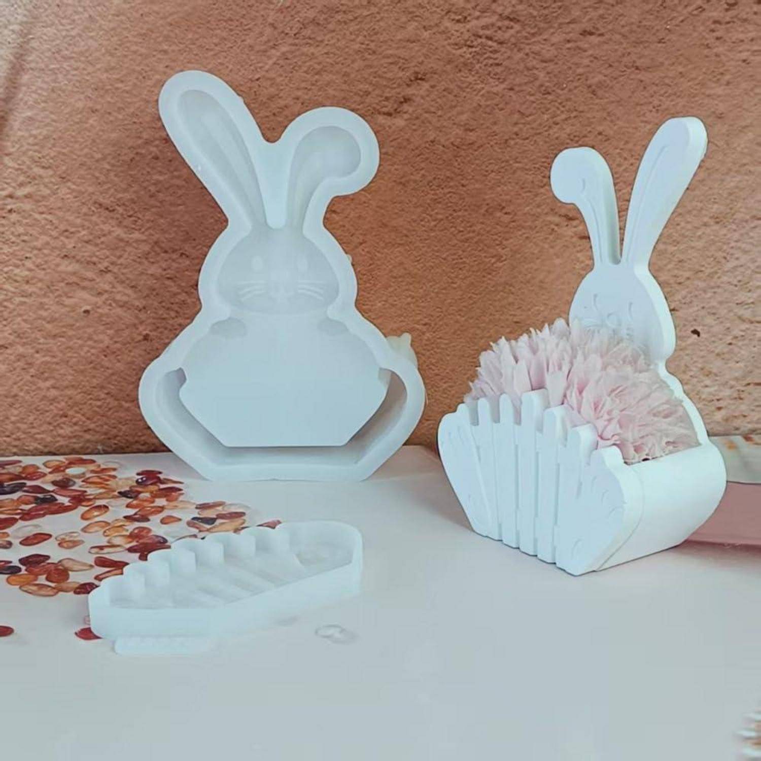 Weiße kaninchenförmige Silikonformen und passende Behälter auf einem Tisch, einer leer stehend und einer mit rosa Blumen gefüllt, neben einem kleinen Tablett.