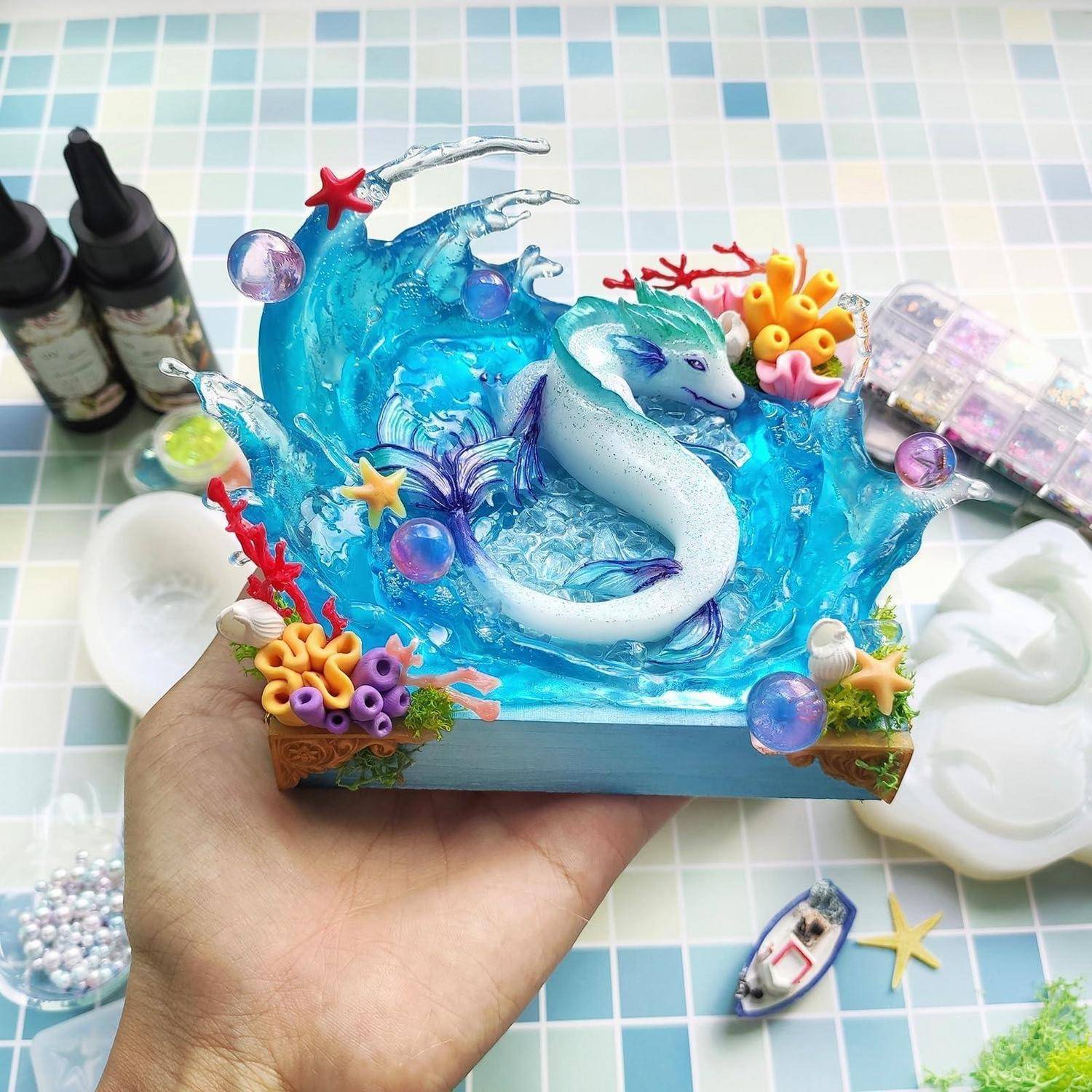 Hand, die ein kleines Harz-Ozean-Diorama mit einem weißen Seepferdchen inmitten blauer Wellen, Korallen und Muscheln hält.