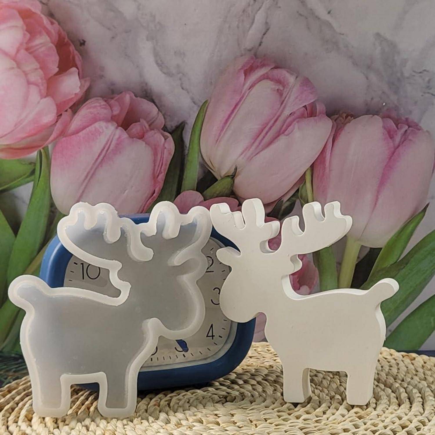 Zwei weiße rentierförmige Kerzenhalter oder Ornamente auf einer gewebten Matte, mit einer blauen Analoguhr und rosa Tulpenhintergrund dahinter.