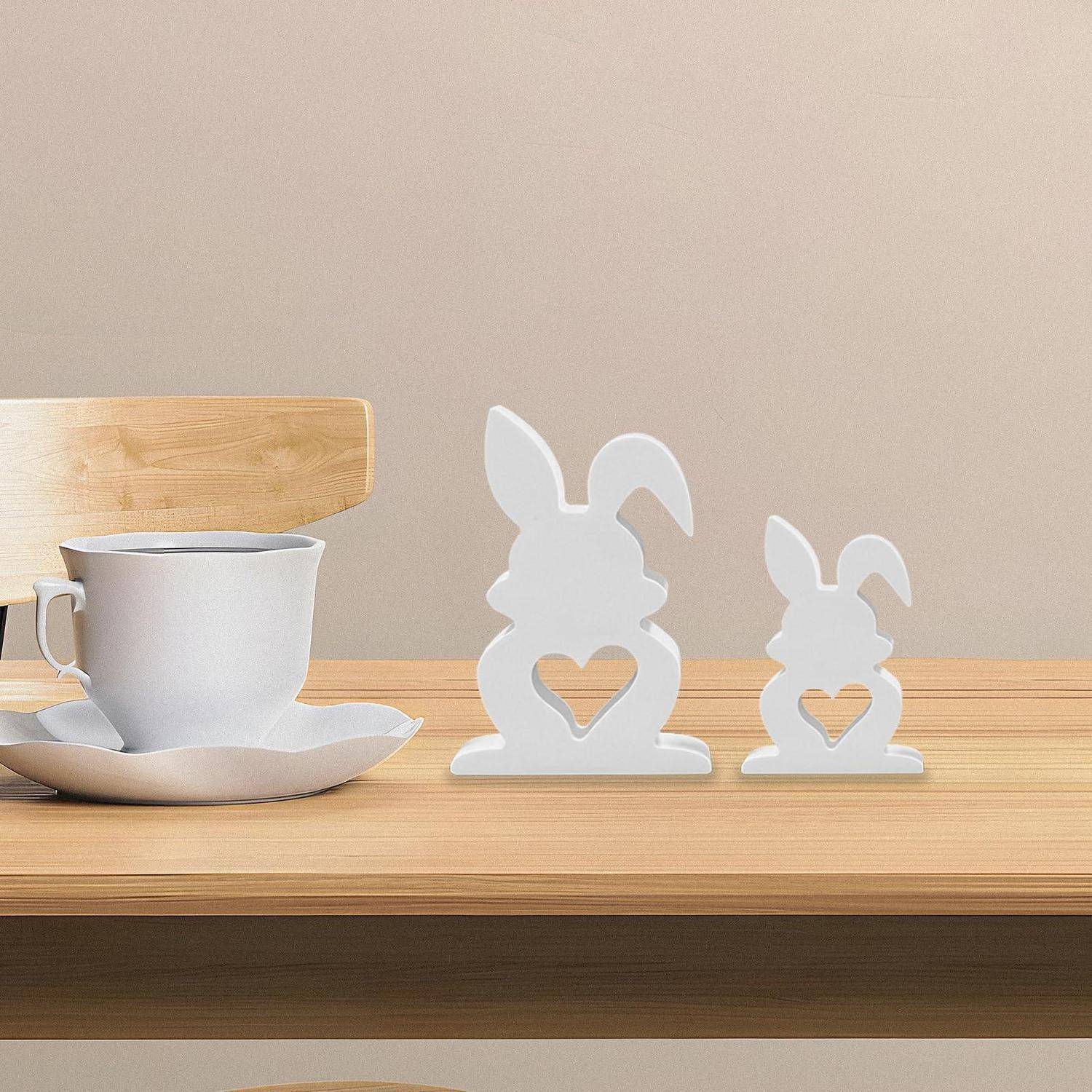 Zwei weiße Kaninchensilhouetten-Dekorationen auf einem Holzregal neben einer weißen Teetasse und Untertasse, mit einem Holzstuhl im Hintergrund.