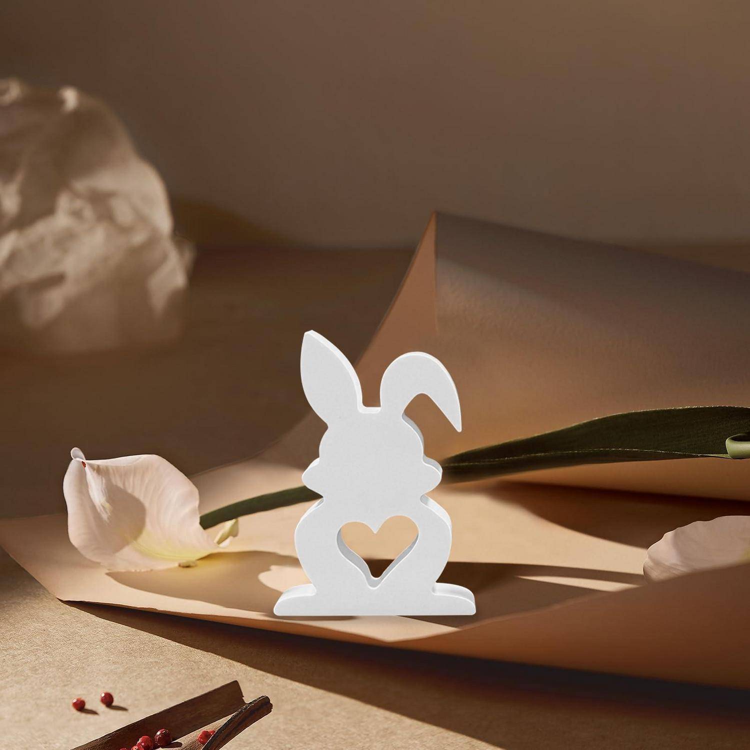 Weiße Hasensilhouette mit Herzausschnitt steht auf einer beigen Papierfläche neben Blütenblättern in warmem Licht, wie eine Osterdekoration.