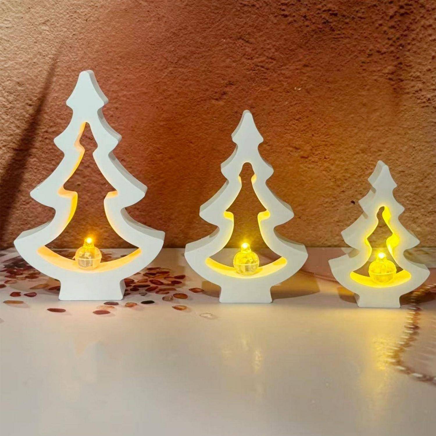 Drei weiße Weihnachtsbaumdekorationen mit warm leuchtenden Lichtern stehen auf einer weißen Oberfläche vor einer strukturierten braunen Wand.