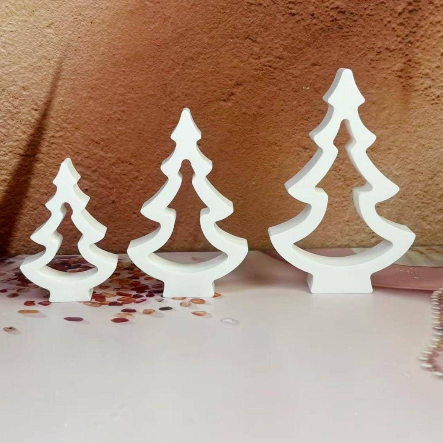 Drei weiße dekorative Weihnachtsbaum-Ausschnitte in aufsteigenden Größen stehen auf einer Tischplatte vor einer strukturierten braunen Wand.
