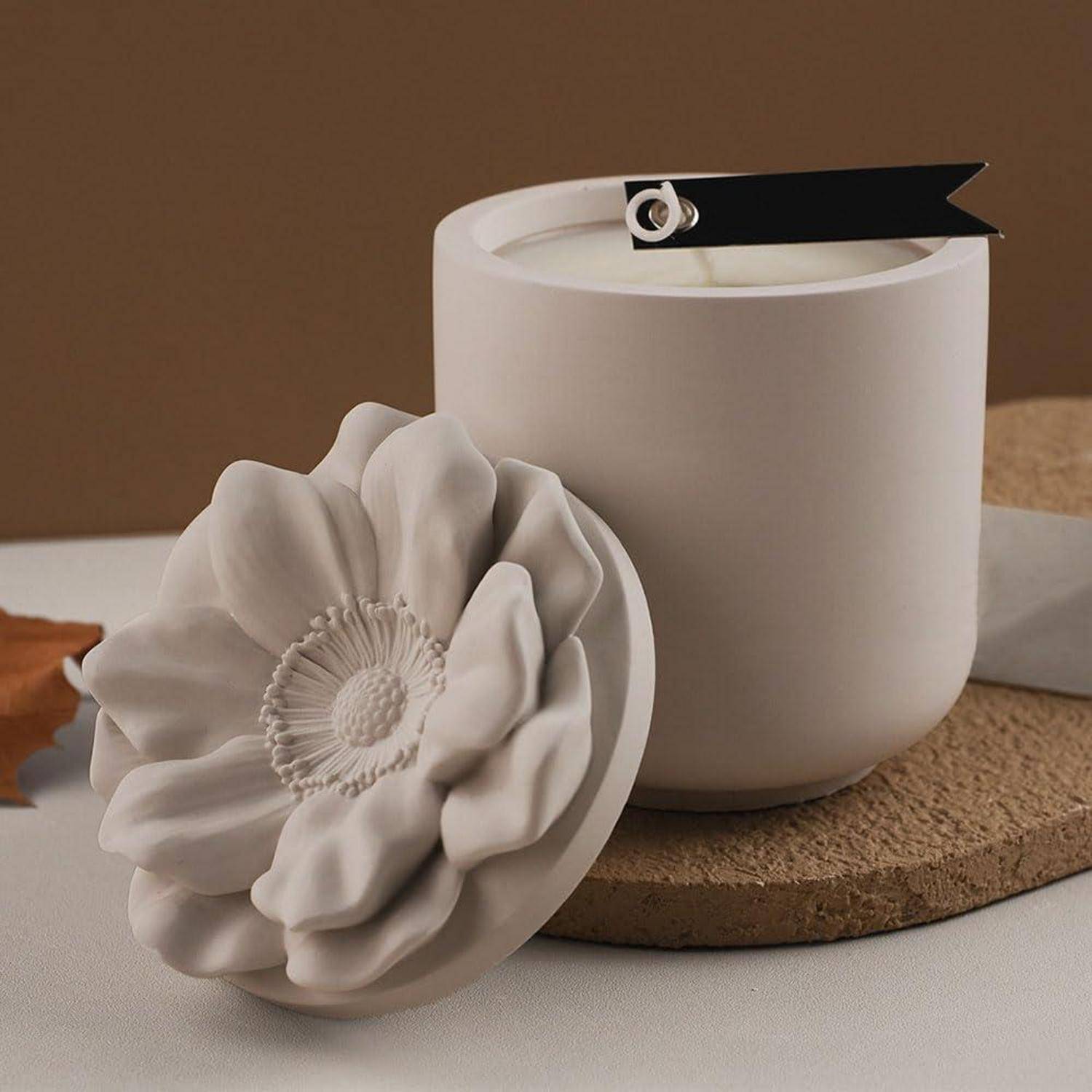 Weiße Kerze in einem matter Keramikhalter mit großer weißer Blumendekoration, stilisiert auf einem Korkuntersetzer vor braunem Hintergrund.