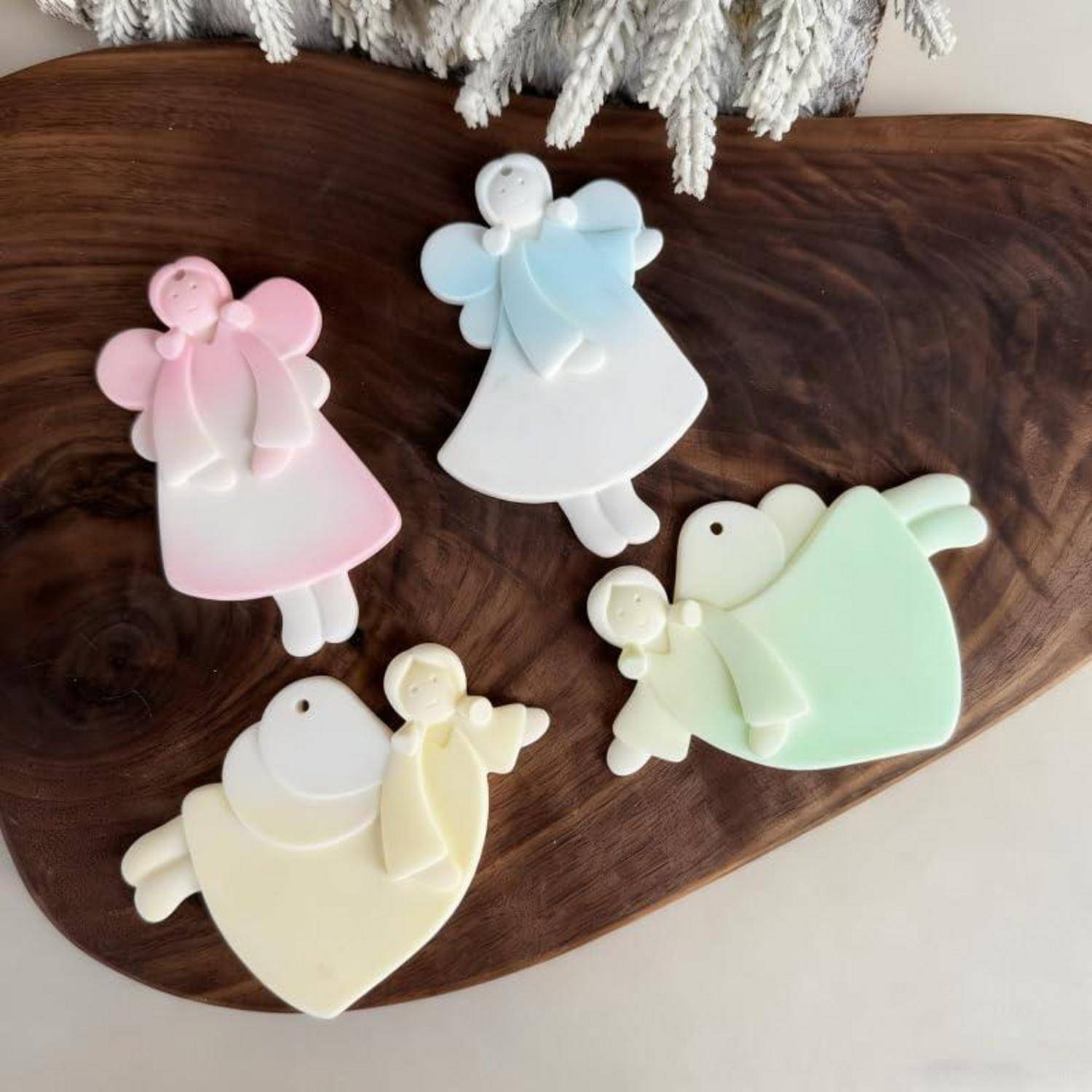 Vier pastellfarbene engelsförmige Ornamente in Rosa, Blau, Weiß und Grün auf einem Holzbrett angeordnet.