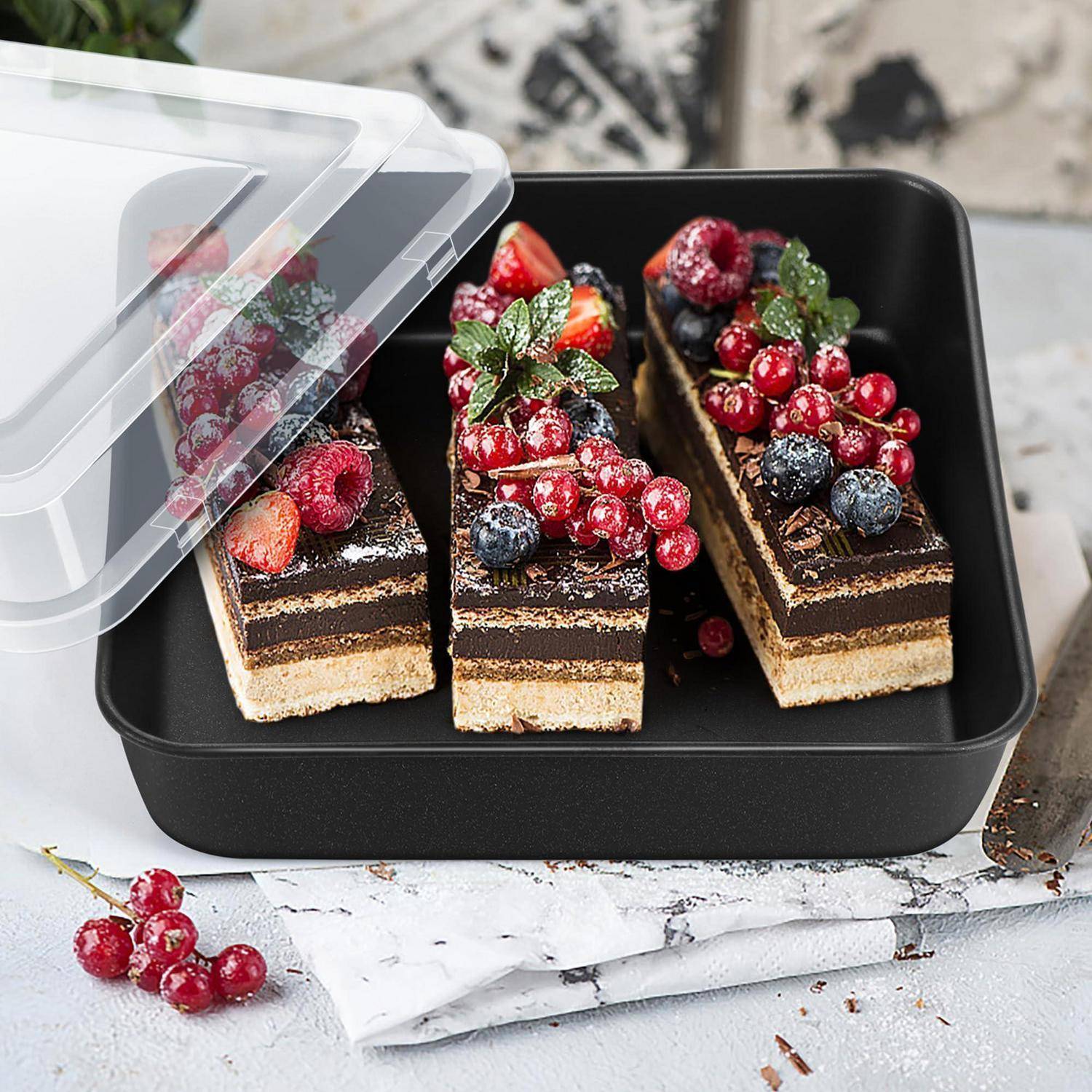 Drei Schichten aus Schokoladenkuchen mit gemischten Beeren belegt in einem schwarzen Behälter mit transparentem Deckel, der teilweise offen ist.