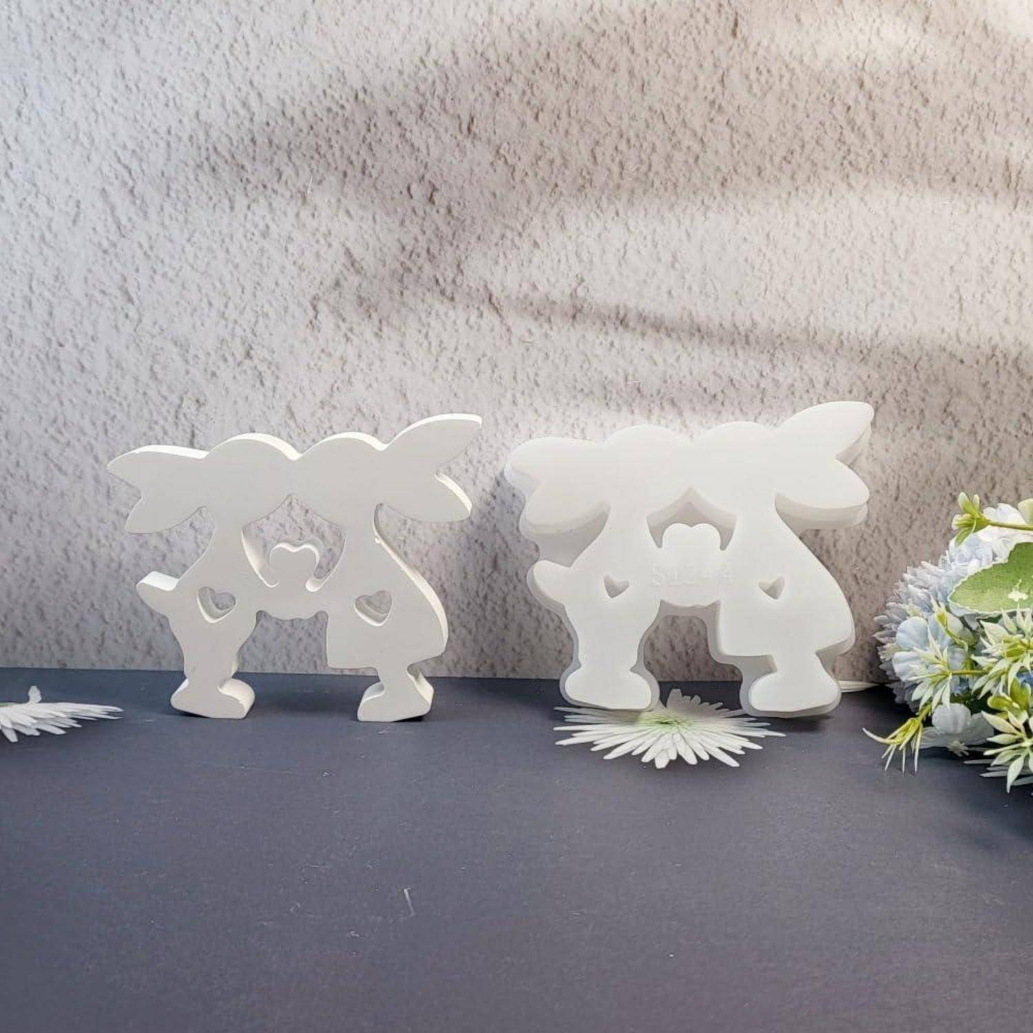 Zwei weiße dekorative Figuren in Form von stilisierten Tieren oder Ornamenten stehen auf einer dunklen Oberfläche, mit einer Blumenanordnung rechts.