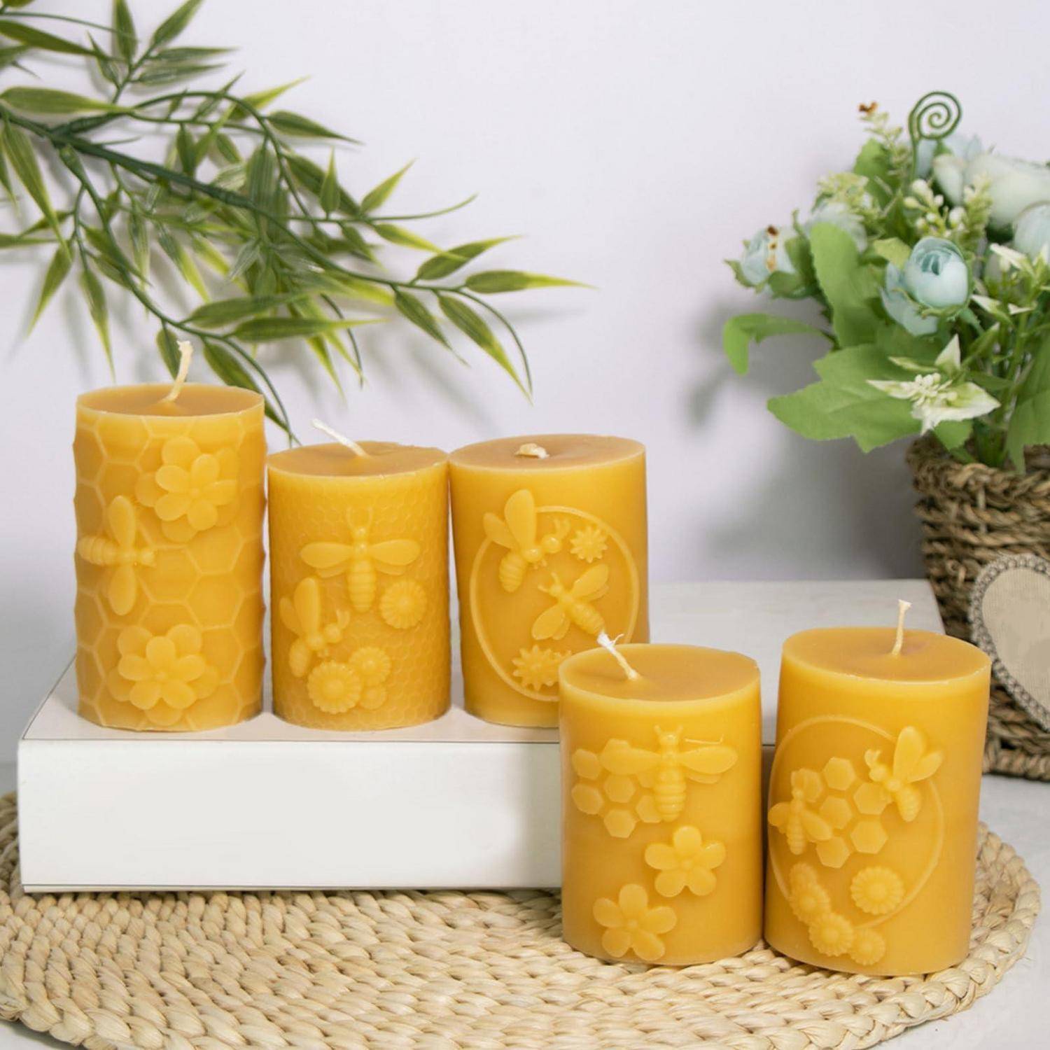 Fünf gelbe dekorative Säulenkerzen mit Blumen- und Bienenmustern, arrangiert auf einer gewebten Matte neben Pflanzen und einem Blumenstrauß.