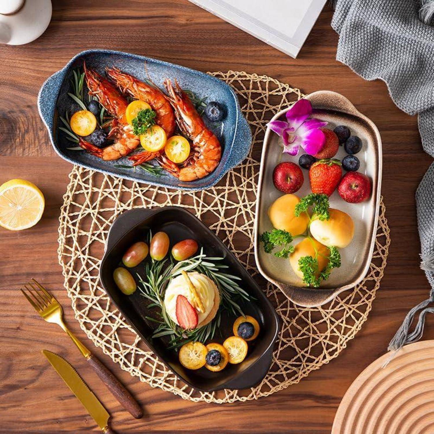 Draufsicht auf einen Holztisch mit drei angerichteten Gerichten: gegrillte Garnelen, Obst mit Orchidee und ein Dessert, angeordnet auf einer geflochtenen Platzdecke.