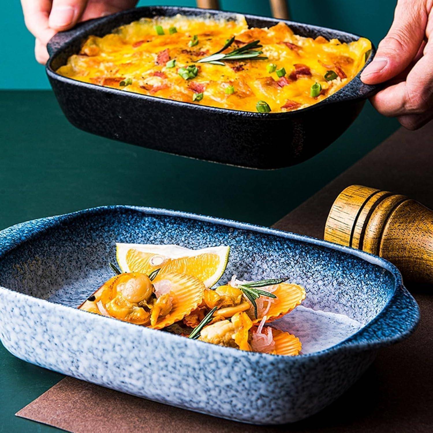 Hände halten eine schwarze Auflaufform mit gebackenem Auflauf über eine blaugesprenkelte Schüssel mit Meeresfrüchte-Pasta, angerichtet auf einem Tisch.