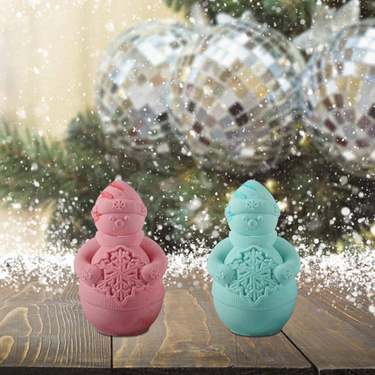 Zwei kleine Schneemannfiguren, eine rosa und eine mintgrün, stehen auf einer Holzoberfläche mit fallendem Schnee und unscharfen Weihnachtskugeln im Hintergrund.