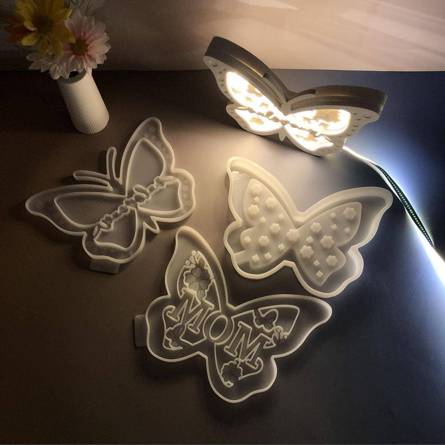 Drei schmetterlingsförmige LED-Wandleuchten auf einer dunklen Oberfläche, einschließlich einer leuchtenden Schmetterlingslampe und einer mit "MOM""-Text.
