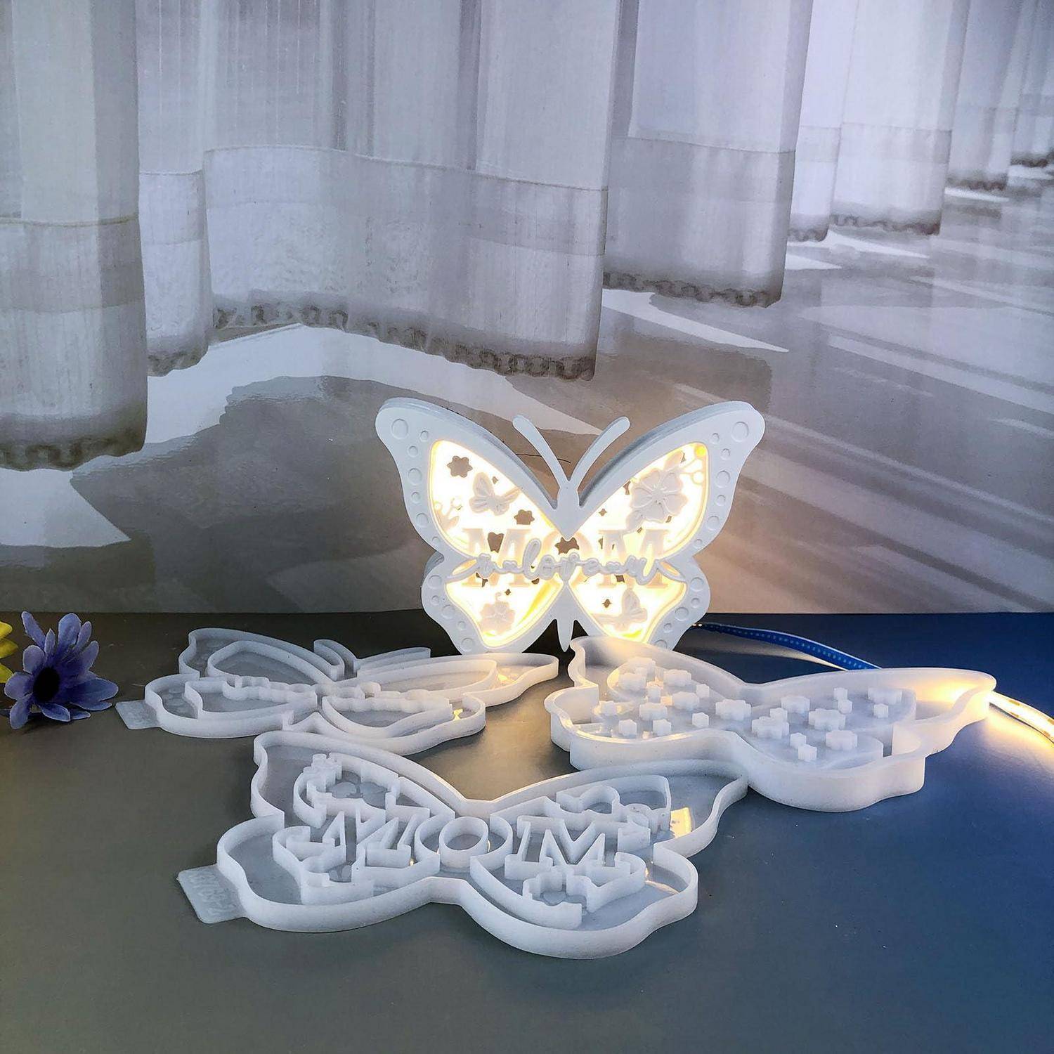 Aus dem 3D-Drucker gefertigtes, schmetterlingsförmiges leuchtendes Schild mit strahlenden warmen Lichtern, umgeben von weißen dekorativen Ausschnittpapieren auf einem Tisch in der Nähe von transparenten Gardinen.