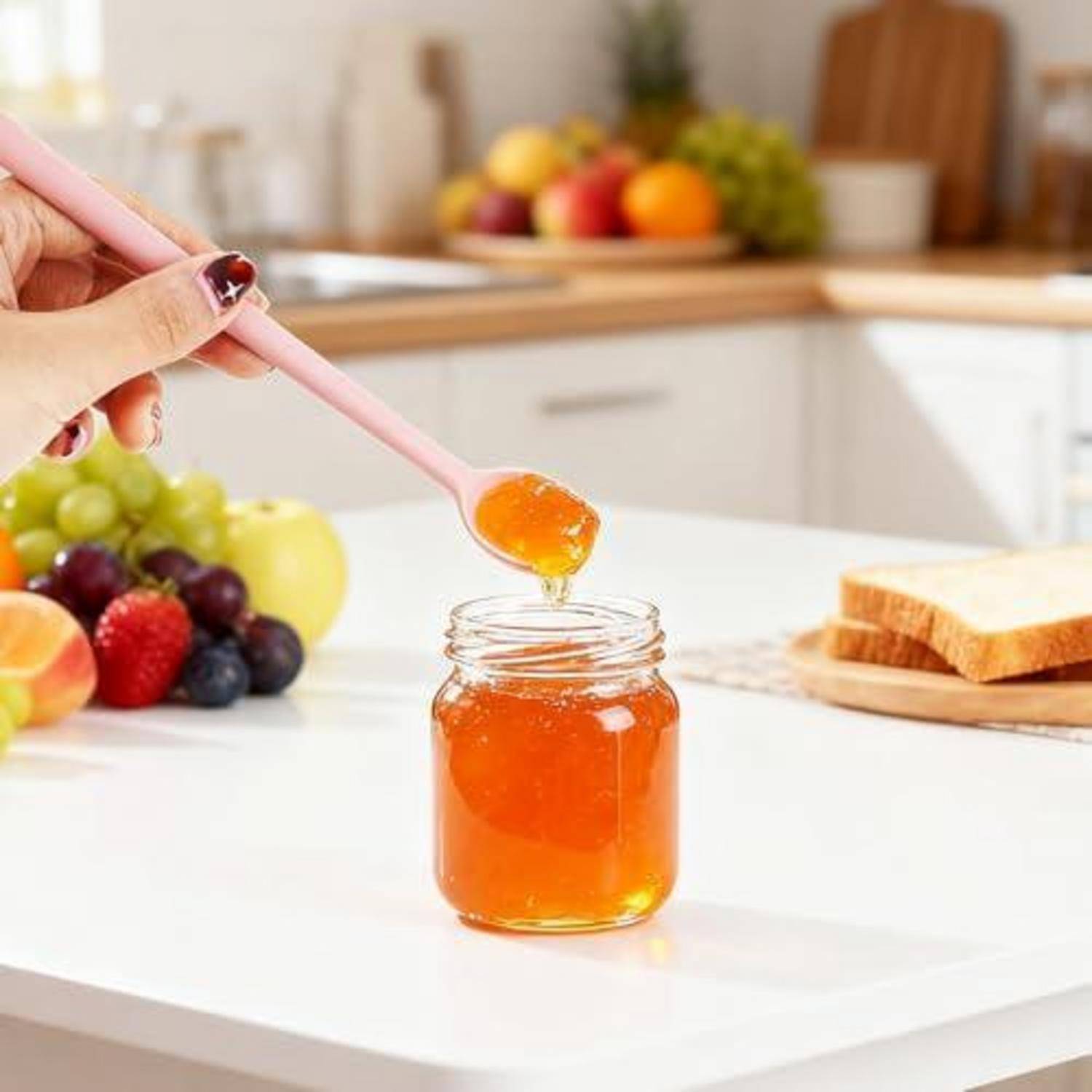 Hand, die orangefarbene Marmelade auf einen Löffel in ein Glasgefäß in einer hellen Küche legt, mit Obst und Brot im Hintergrund.
