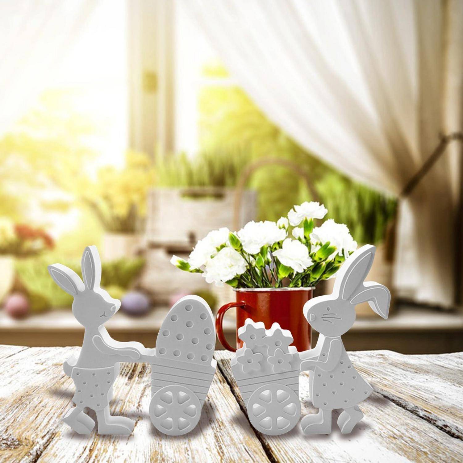 Zwei graue Kaninchenfiguren und zwei Ostereier auf einem rustikalen Tisch, mit weißen Blumen in einer roten Tasse im Hintergrund.