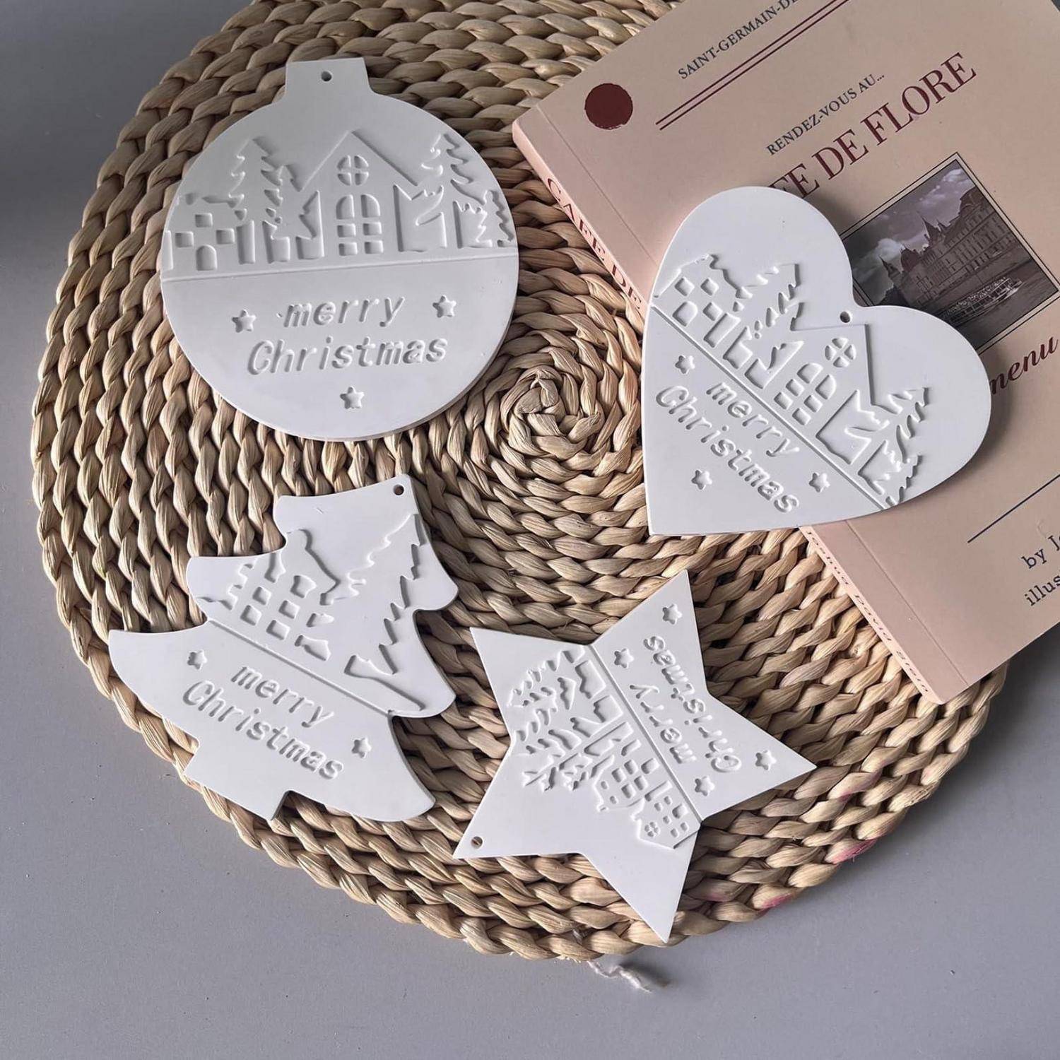 Vier weiße geprägte „Frohe Weihnachten"-Ornamente in runder, Herz-, Baum- und Sternform auf einer geflochtenen Platzdecke neben einem rosa Magazin.