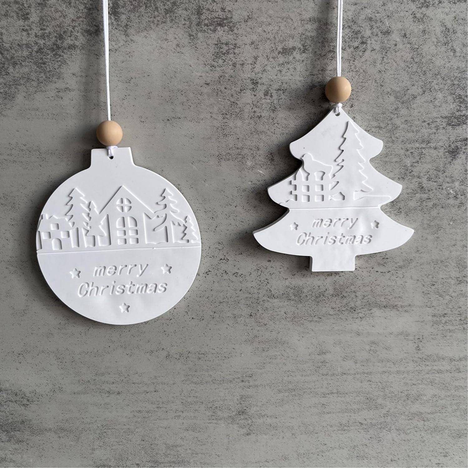 Zwei weiße hängende Weihnachtsverzierungen, eine rund und eine baumförmig, jeweils mit "frohe Weihnachten"" und Winterdorfdesigns geprägt.