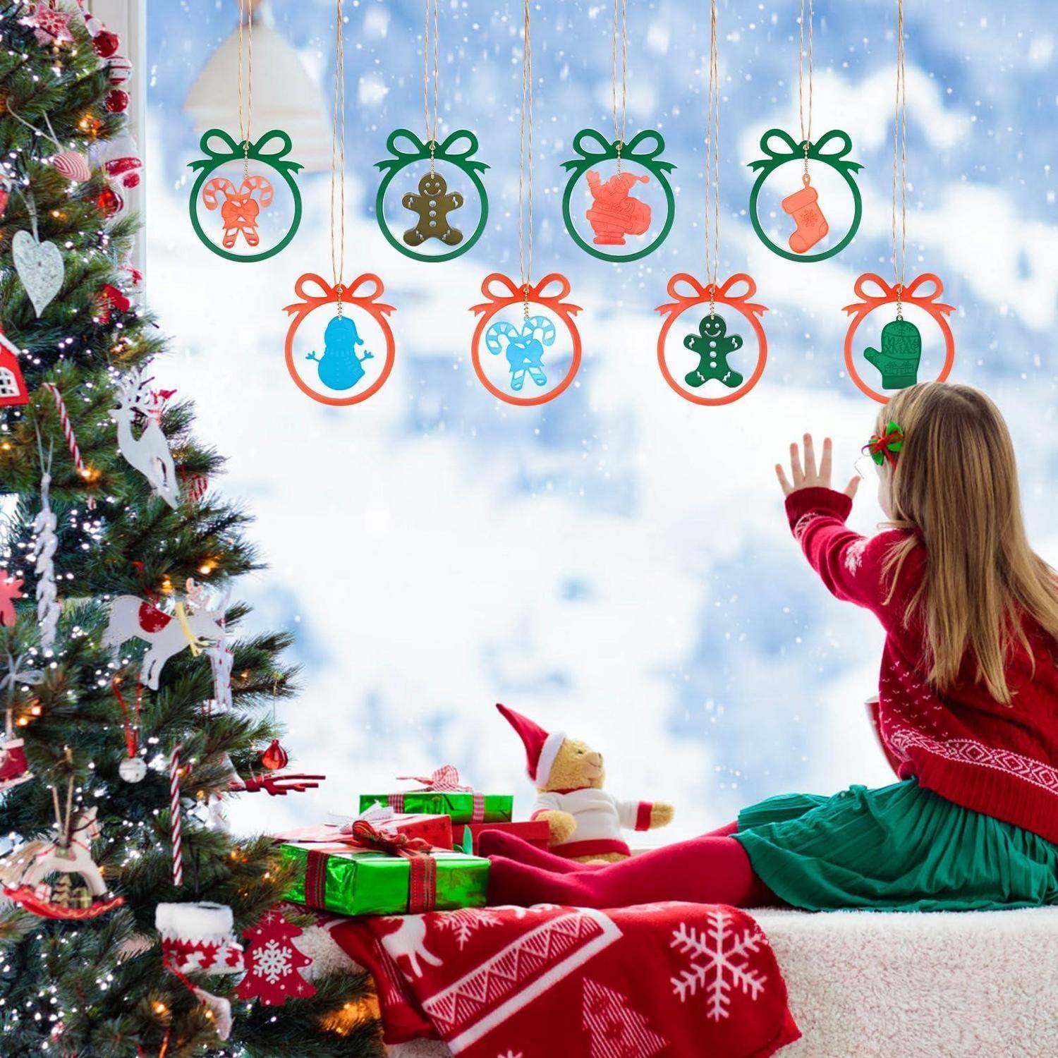 Mädchen in einem roten Pullover und grünem Rock sitzt neben einem geschmückten Weihnachtsbaum und winkt zu hängenden Christbaumschmuckdekorationen mit schneebedecktem Fenster im Hintergrund.