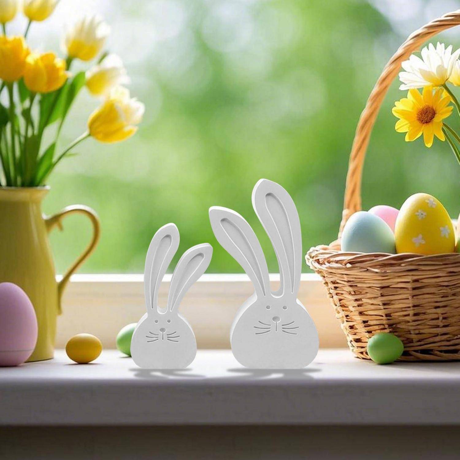 Zwei weiße Hasendekrationen auf einer Fensterbank mit pastellfarbenen Ostereiern, Blumen in einer Vase und einem Korb mit dekorierten Eiern.