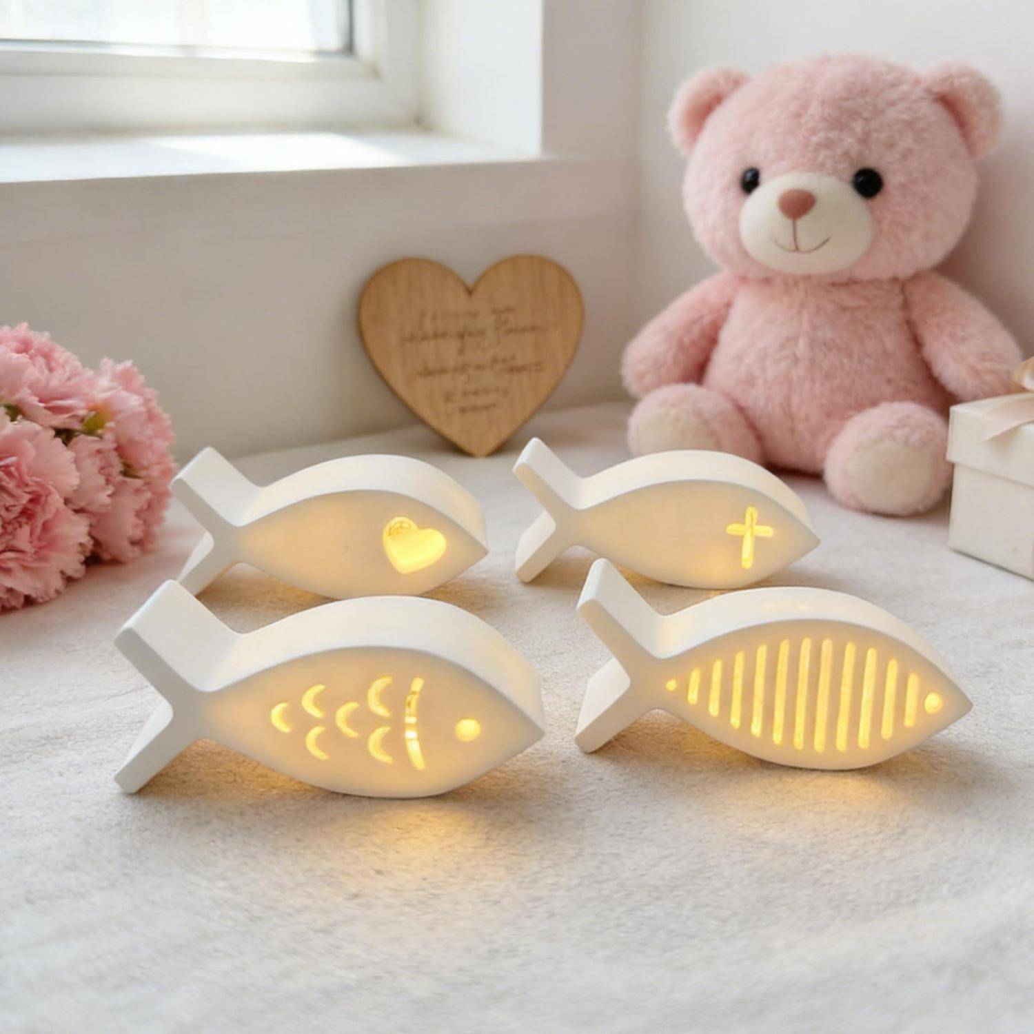 Vier kleine leuchtende fischförmige Nachtlichter auf einer weichen Oberfläche, mit einem Teddybär und einem dekorativen Herz im Hintergrund.