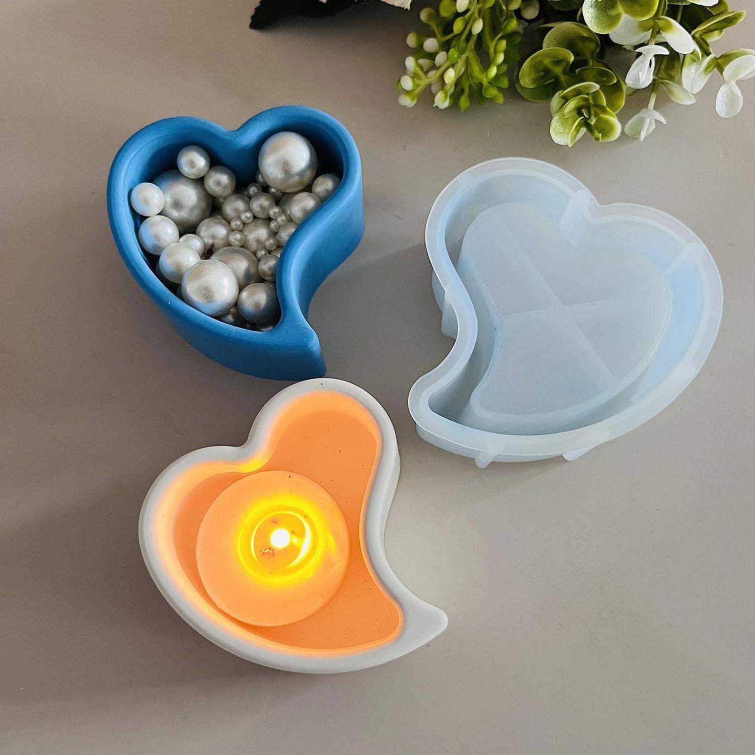 Drei herzförmige Silikonformen auf einem Tisch: eine blaue Form gefüllt mit silbernen Perlen, eine durchscheinende leere Form und eine orangefarbene Form mit einer brennenden Kerze darin.