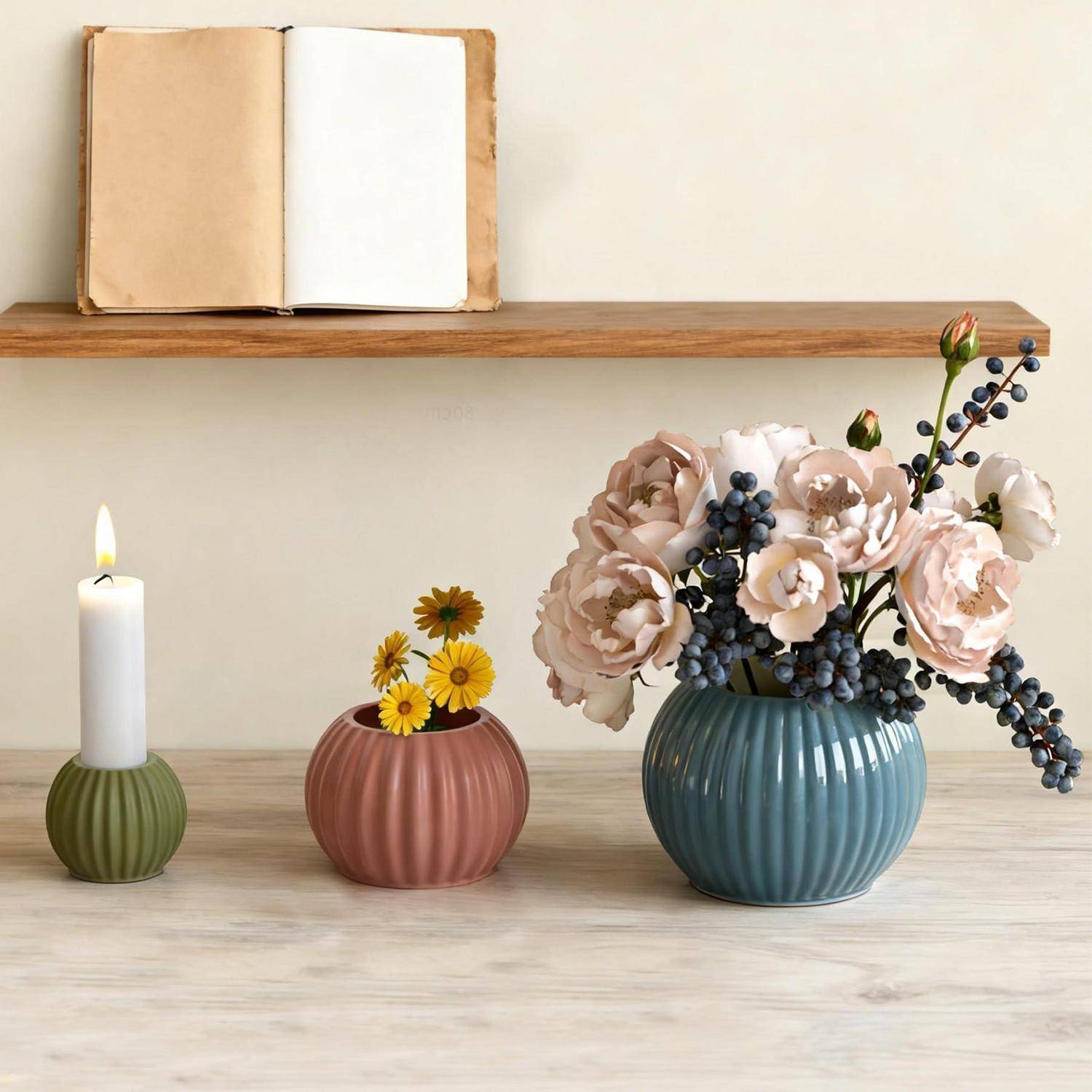 Dekoratives Stillleben auf dem Tisch mit einer brennenden Kerze, einer kleinen Blumenvase und einer größeren blauen Vase mit Blumen unter einem Regal mit einem aufgeschlagenen Buch.