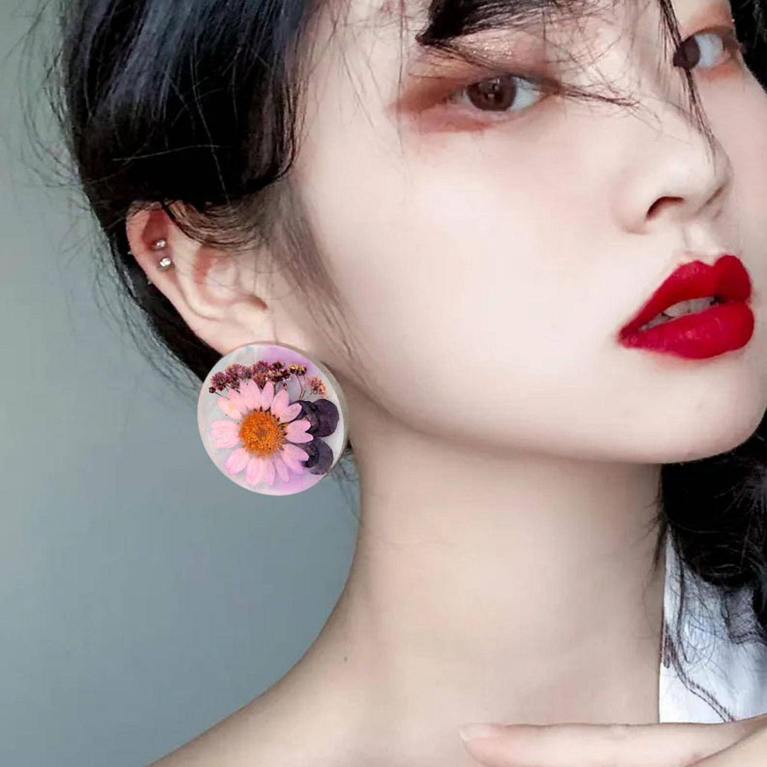 Nahaufnahmeporträt einer Frau mit leuchtend rotem Lippenstift und einem großen Blumenschmuck, nach rechts gewandt vor einem einfachen grauen Hintergrund.