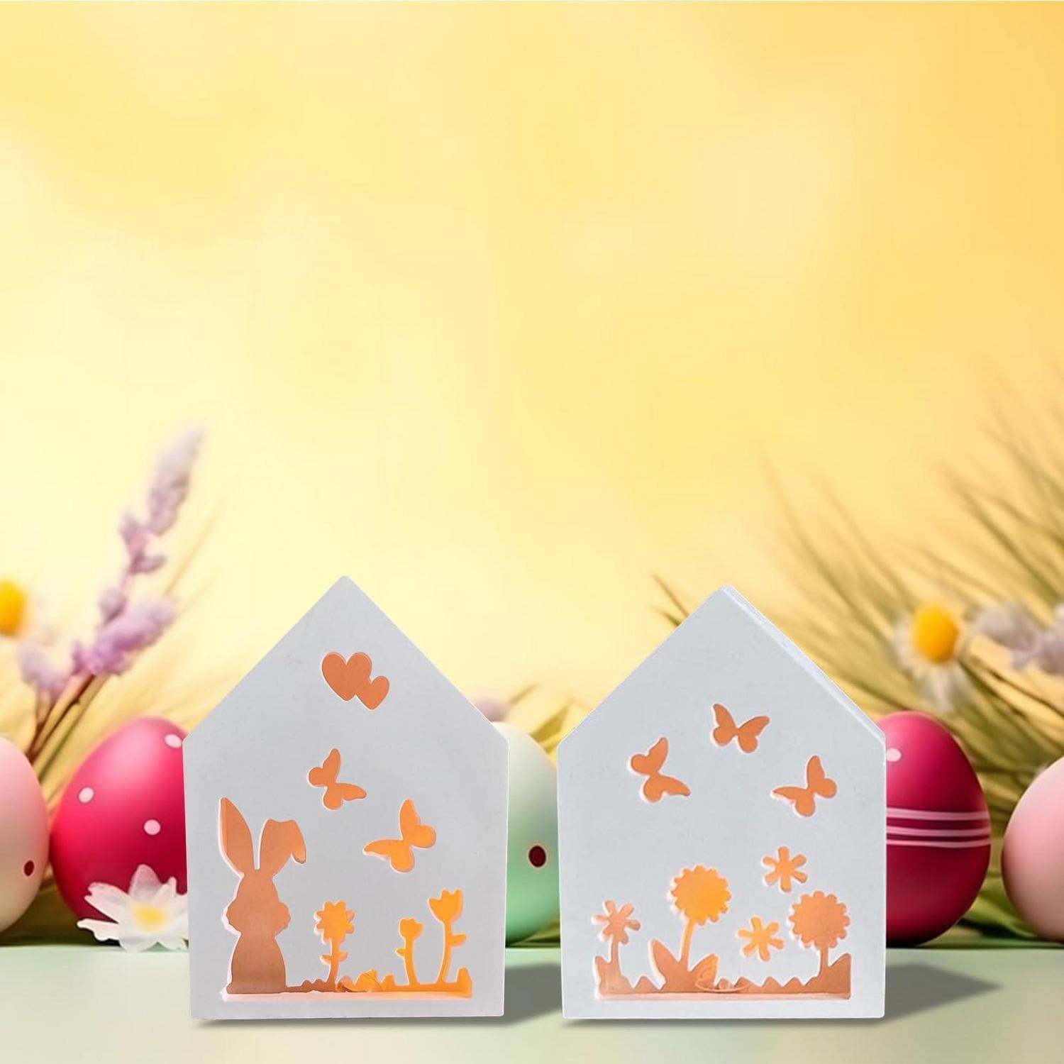 Zwei kleine hausförmige Osterdekoration mit orangefarbigen Hasen- und Schmetterlingsausschnitten, umgeben von pastell bemalten Eiern und Blumen.