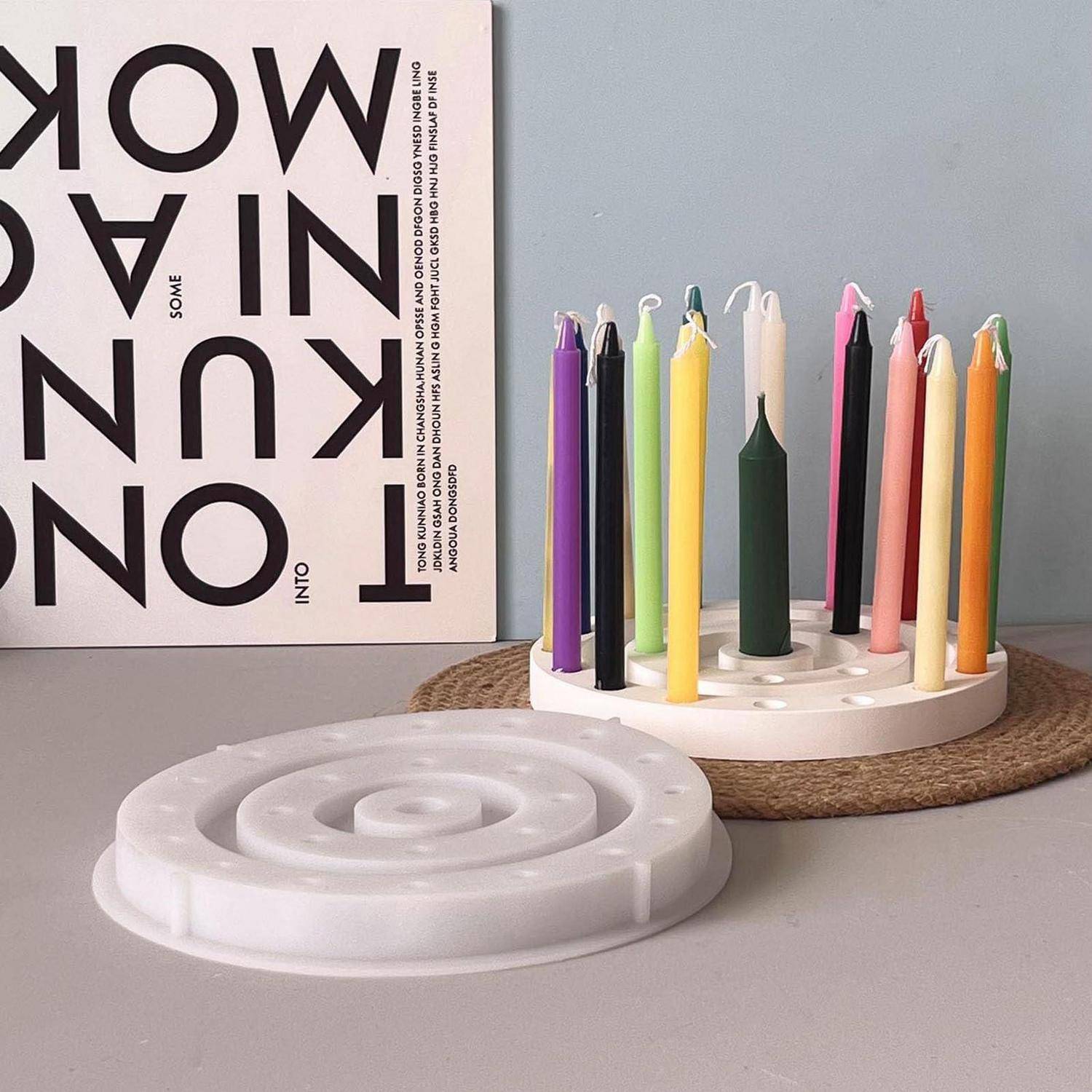 Bunte Stabkerzen stehen in einem weißen rotierenden Kerzenhalter neben einem weißen spiralförmigen Kerzenständer auf einer Tischplatte neben einem Poster.