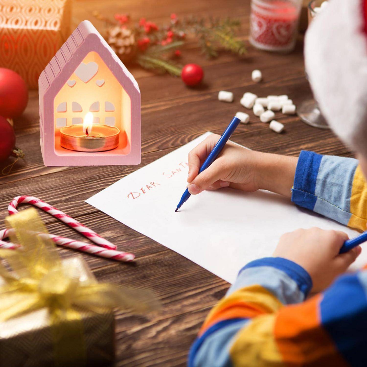 Ein Kind schreibt "LIEBER WEIHNACHTSMANN"" auf eine Karte an einem festlichen Tisch mit einem beleuchteten Kerzenhaus, Zuckerstangen und Weihnachtsdekoration.
