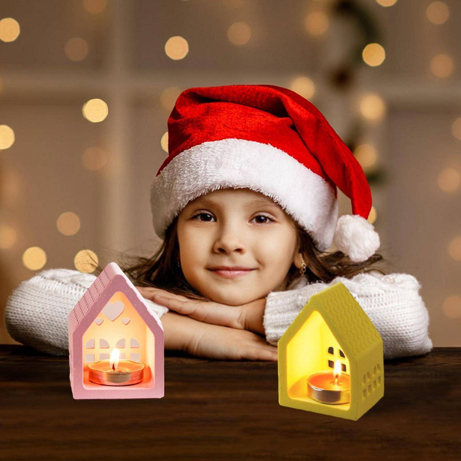 Lachendes Kind in einer Weihnachtsmütze ruht auf einem Tisch neben zwei brennenden hausförmigen Kerzen, mit warmen Weihnachtslichtern im Hintergrund.