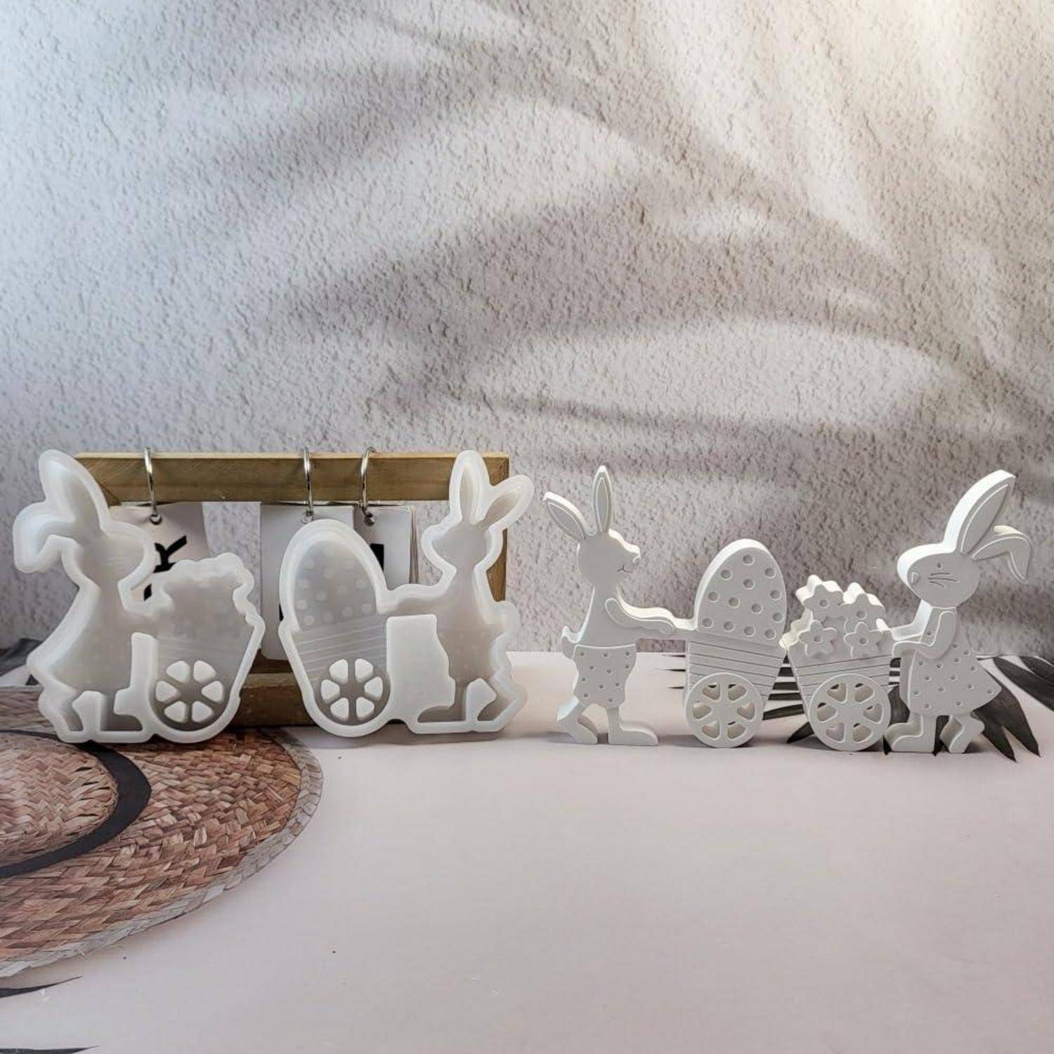 Weiße Osterhasen und Küken aus Holz, die auf einem Tisch vor einer strukturierten Wand arrangiert sind, mit einem Korb- und Eierwagen-Motiv.
