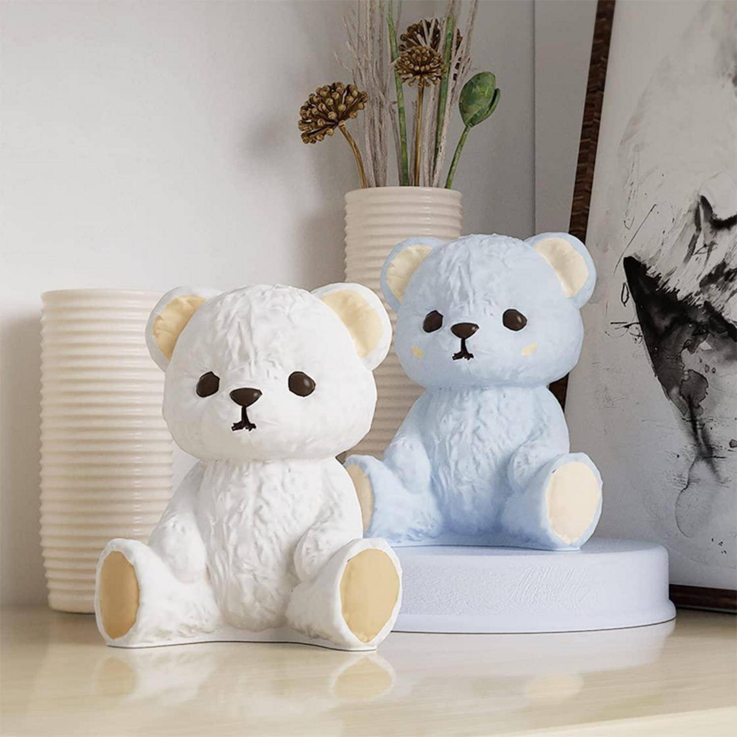 Zwei Teddybär-Figürinen, eine weiße und eine hellblaue, sitzen auf Ausstellungssockeln in einer Heimdekorationsszenerie.