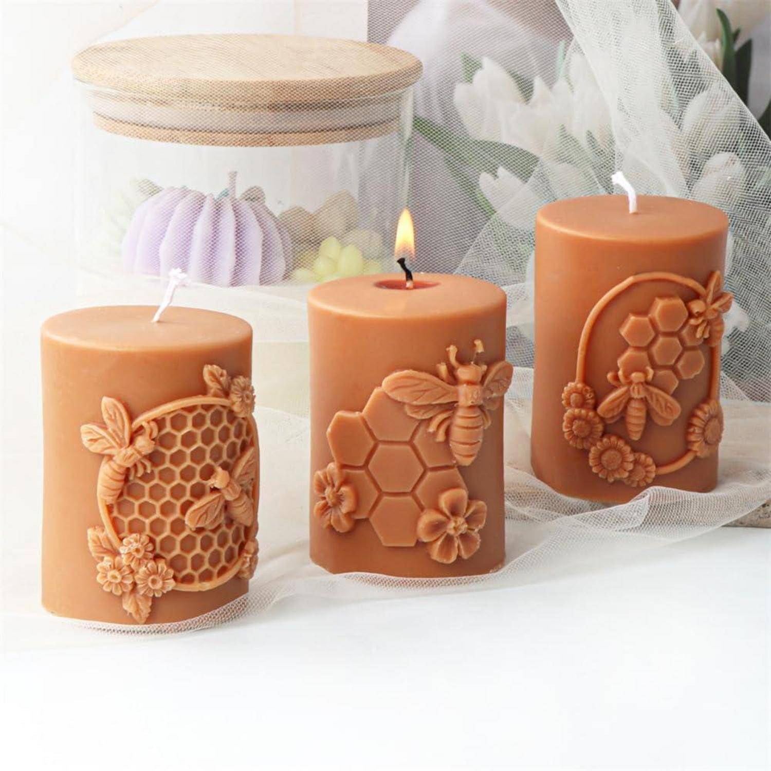 Drei dekorative Bernstein-Kerzenständer mit Wabenmuster- und Bienendesigns, einer davon angezündet, auf einer weißen Oberfläche angeordnet, mit einem Glas mit pastellfarbenen Wachsschmelzen dahinter.